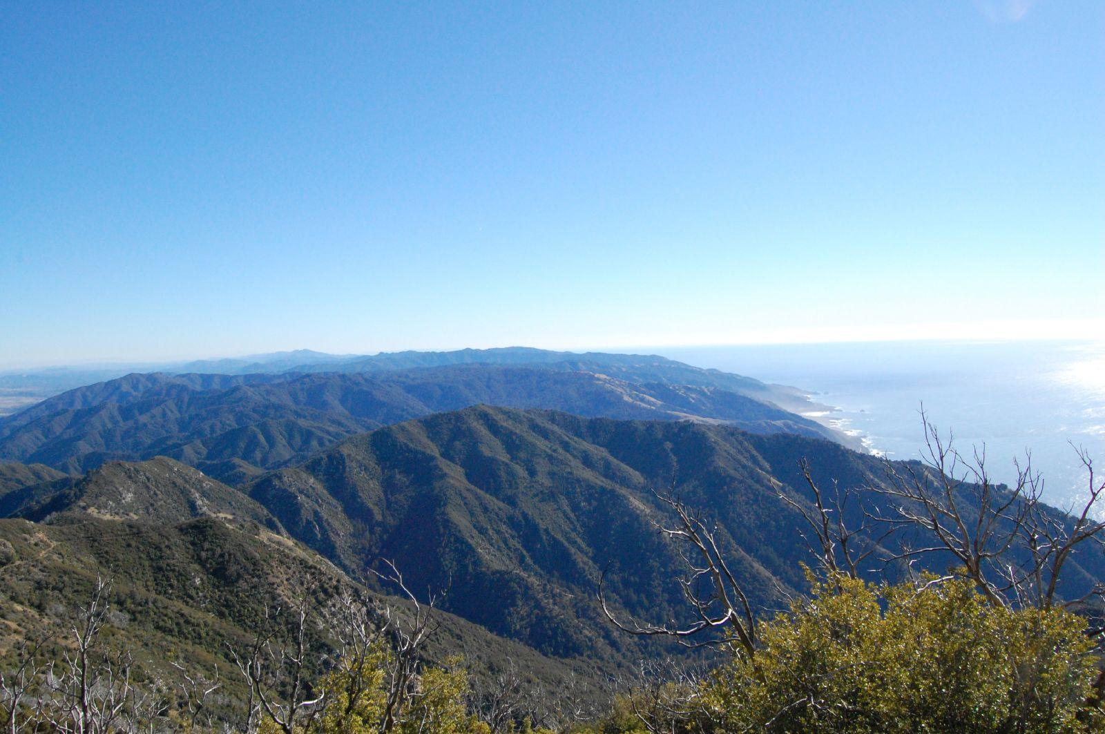Expansive view of green mountains meeting a sparkling ocean under a clear blue sky.
