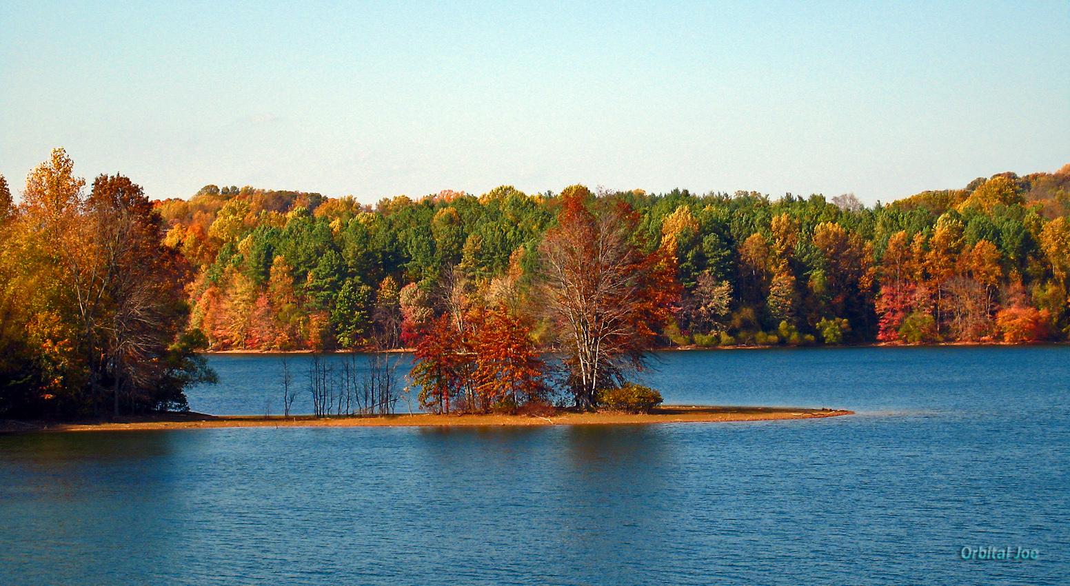 Vibrant autumn trees line a clear blue lake under a bright sky.