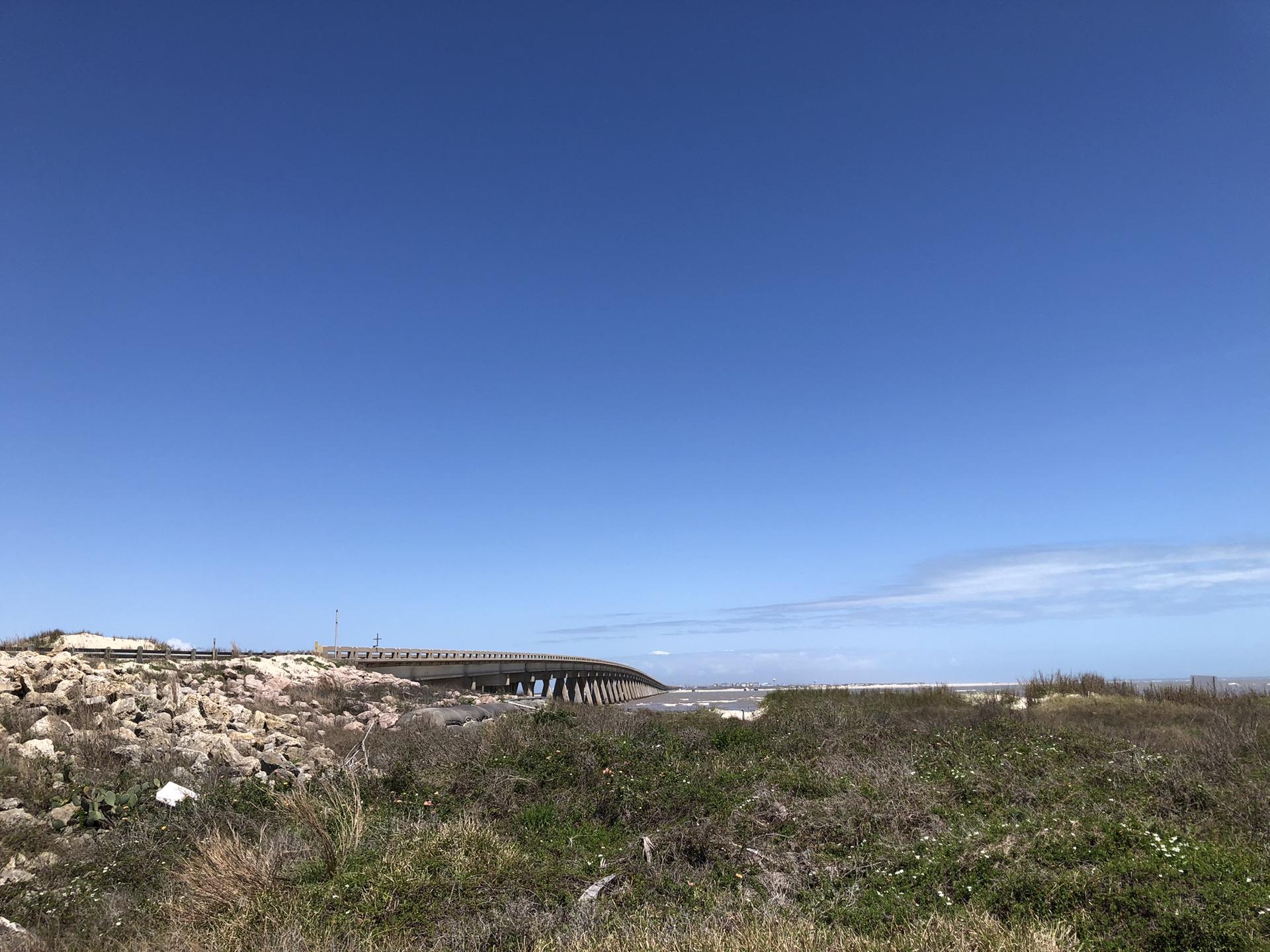 Sunny day with a distant bridge over water and a rocky, grassy foreground.