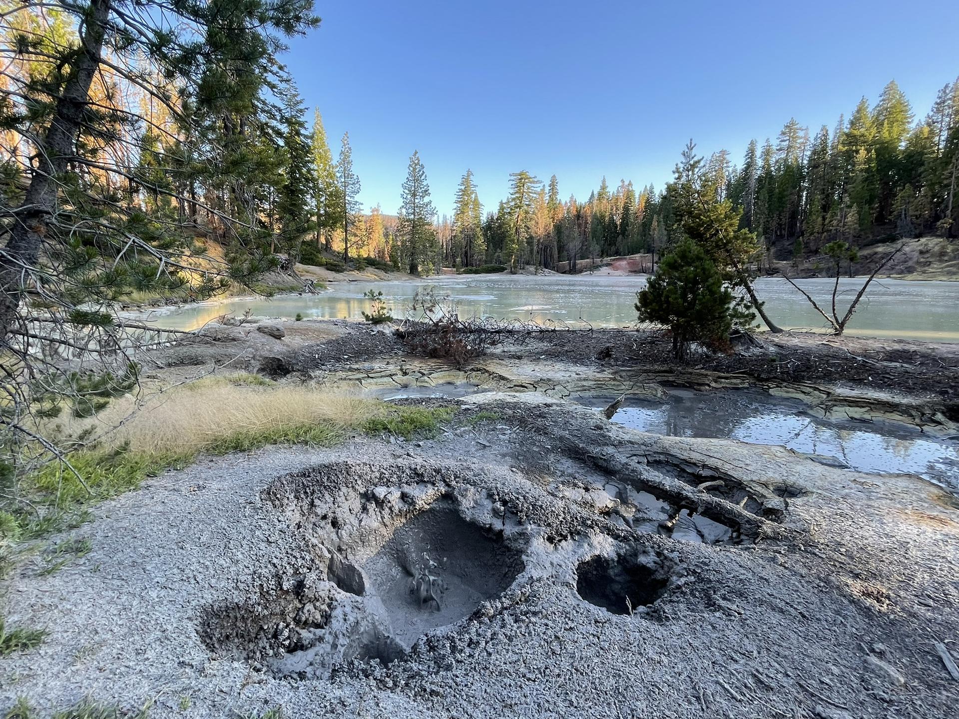 Bubbling mud pots and a light-colored lake surrounded by evergreen trees.