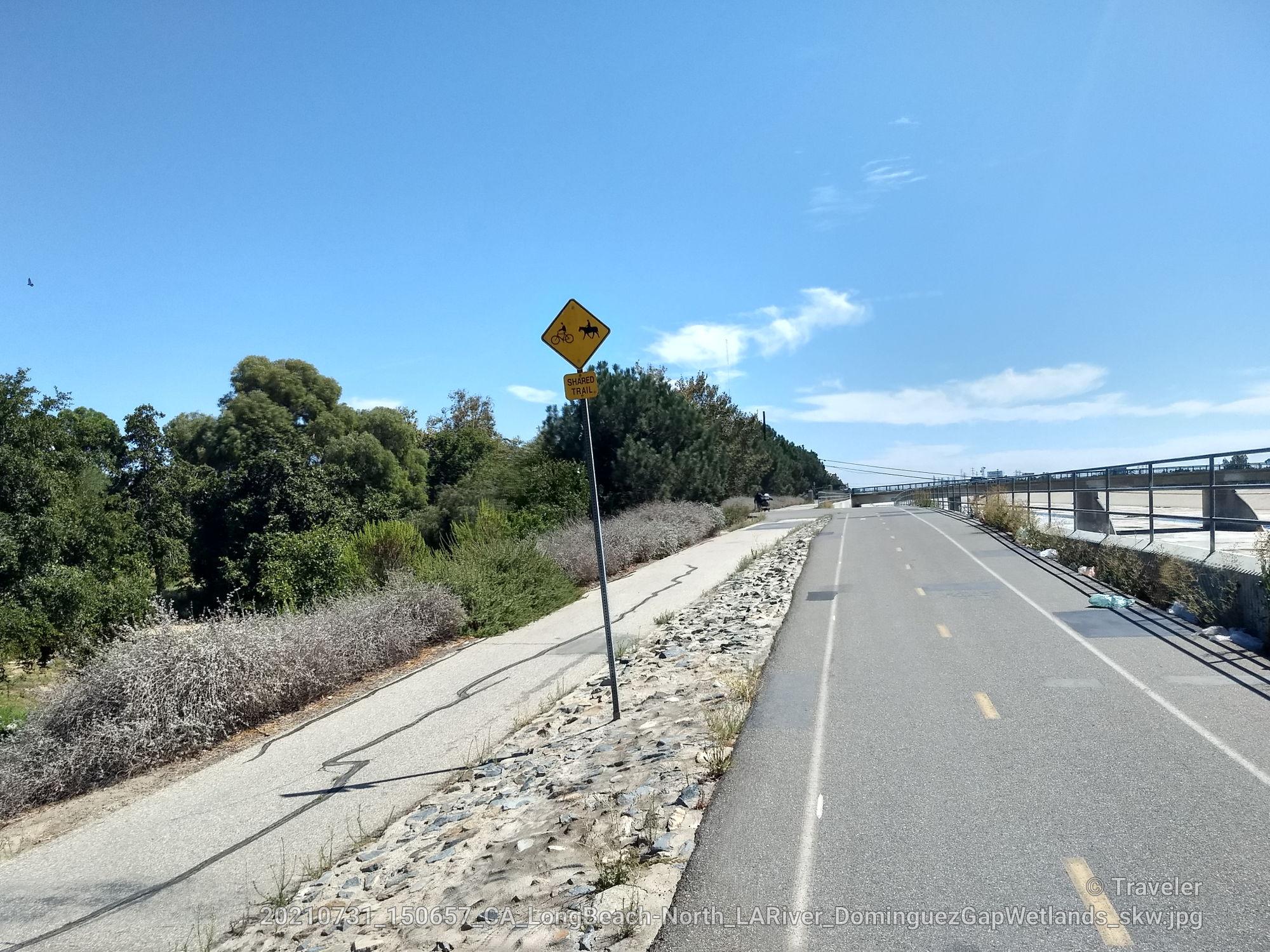 Paved and dirt paths alongside green trees with a warning sign.