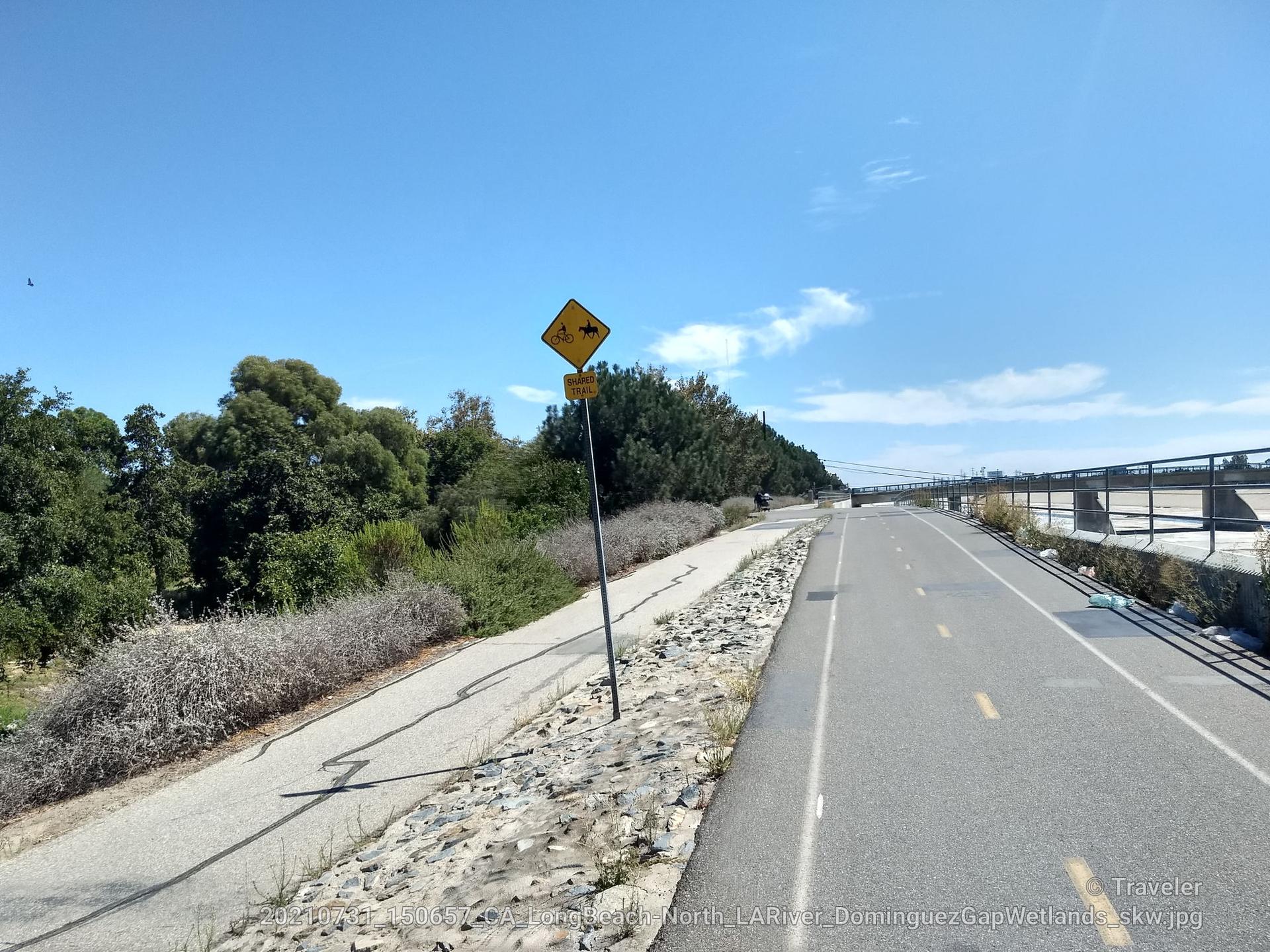 Paved and dirt paths alongside green trees with a warning sign.