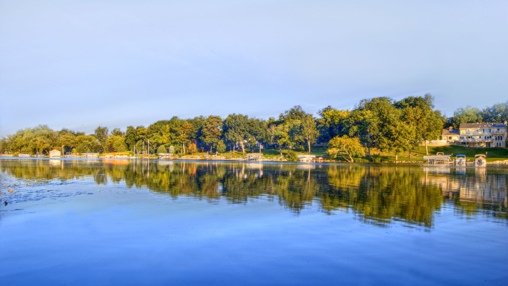 Wide lake reflecting green trees and houses on the distant shore. - Lake Country Trail