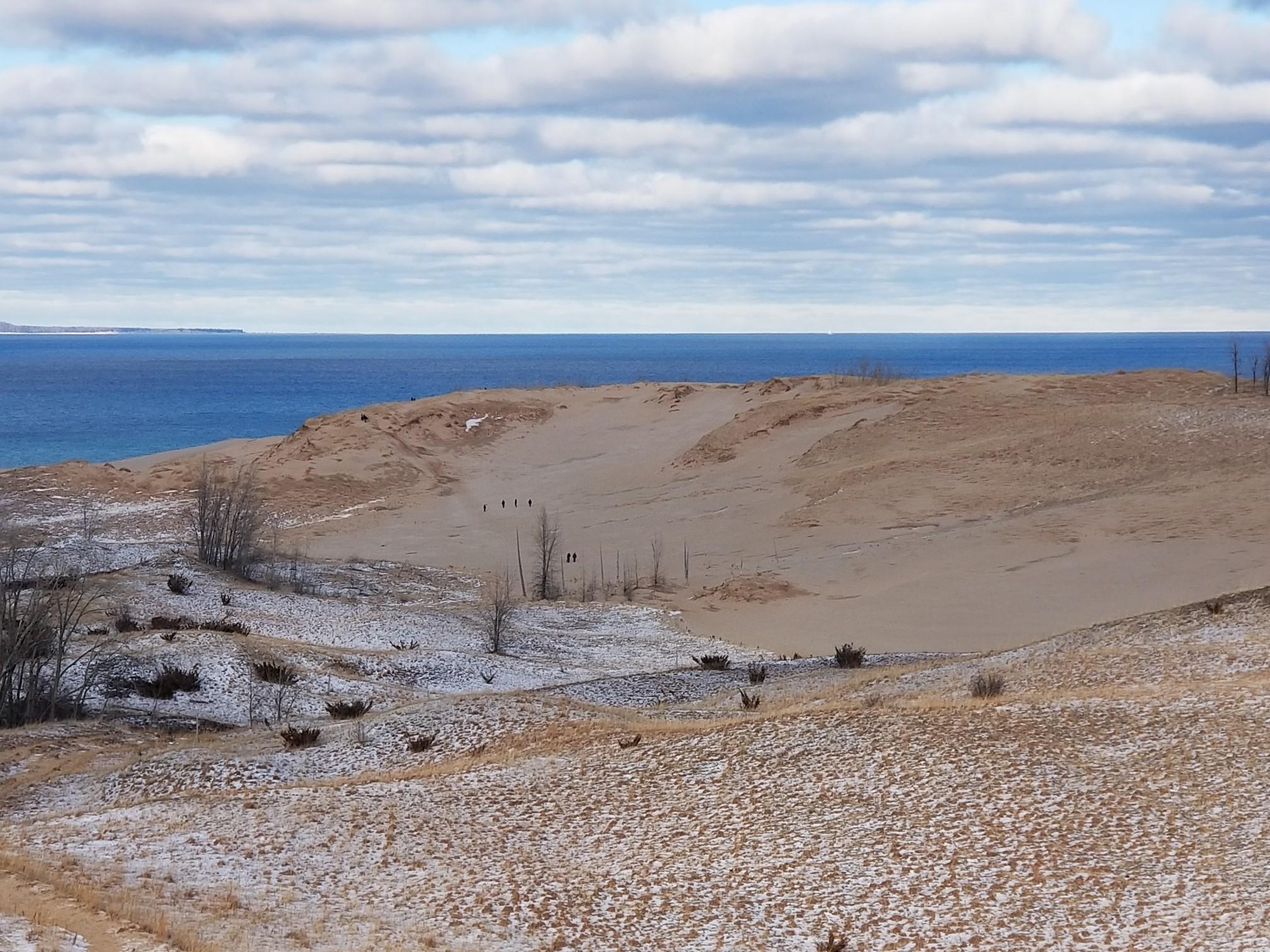 People walk on vast sandy dunes with patches of snow overlooking blue water.