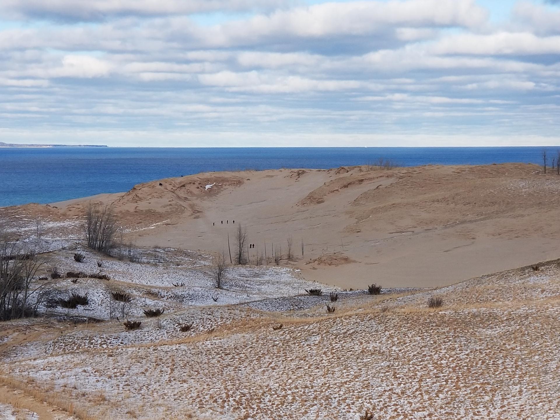 People walk on vast sandy dunes with patches of snow overlooking blue water.