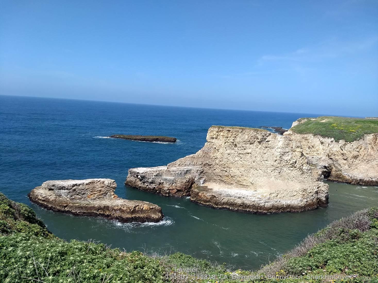 Sunny ocean view with rugged sea stacks, cliffs, and green vegetation.