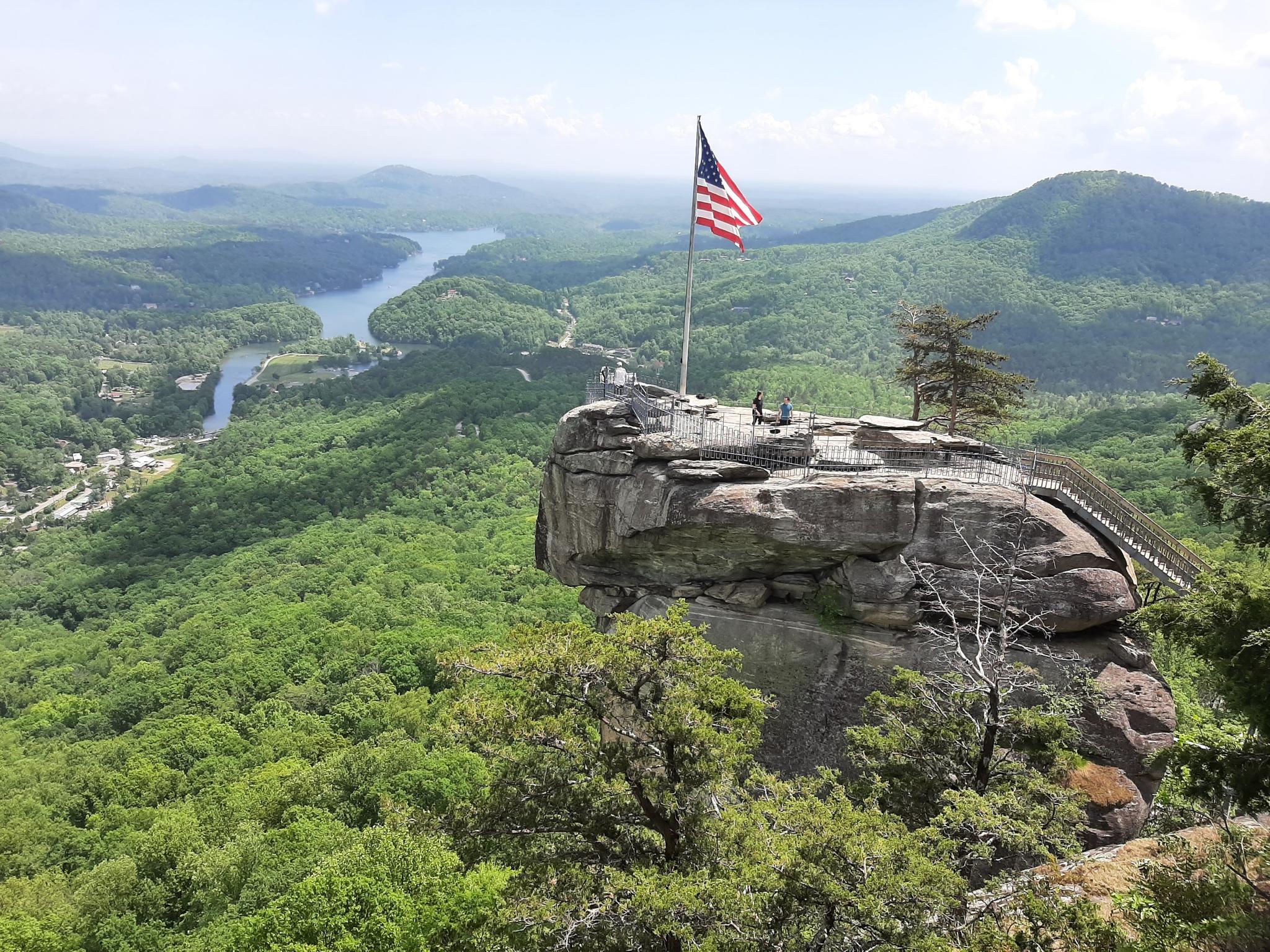 American flag on a rocky peak overlooking a vast green valley and river. Bright daylight, people on peak.