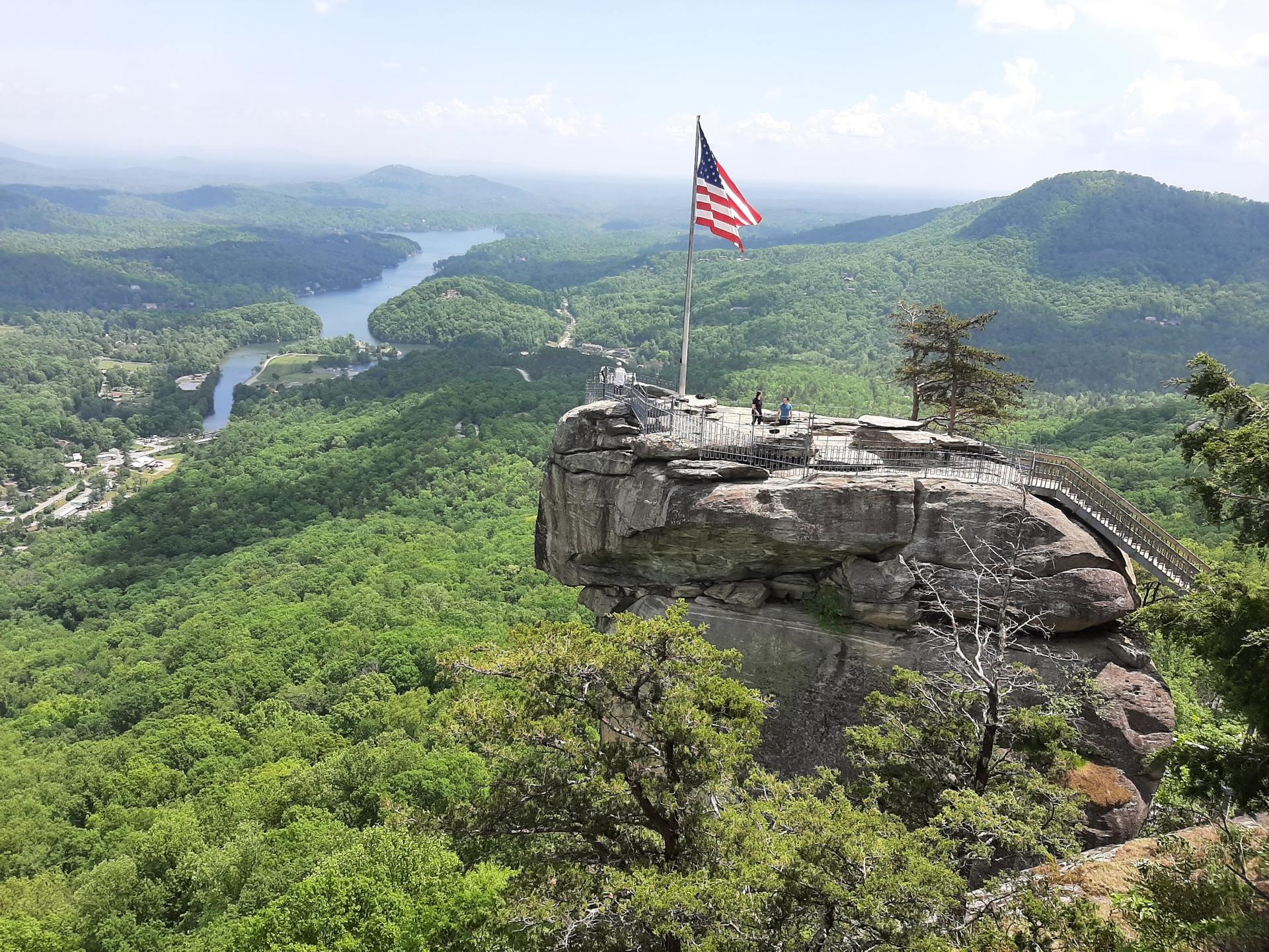 American flag on a rocky peak overlooking a vast green valley and river. Bright daylight, people on peak.