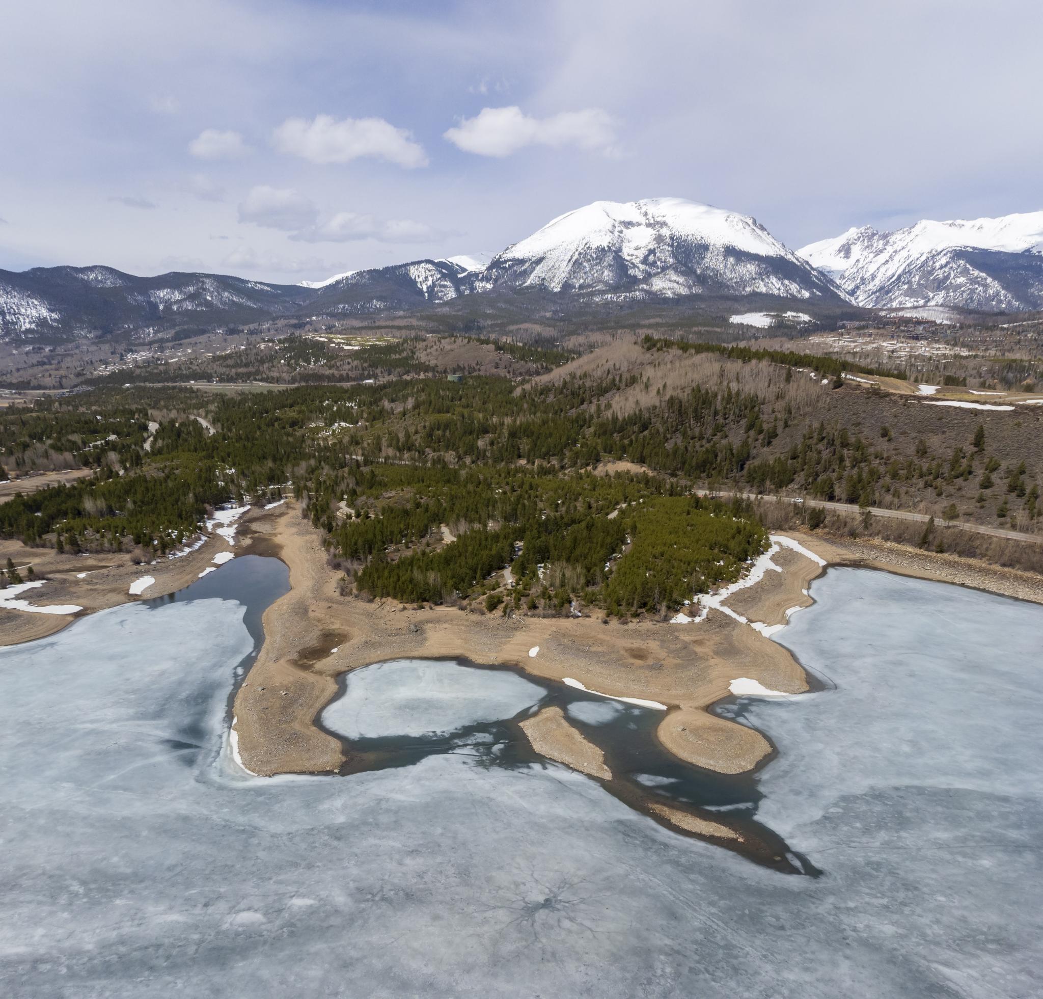 Aerial view of a partially frozen lake, forests, and snow-capped mountains.