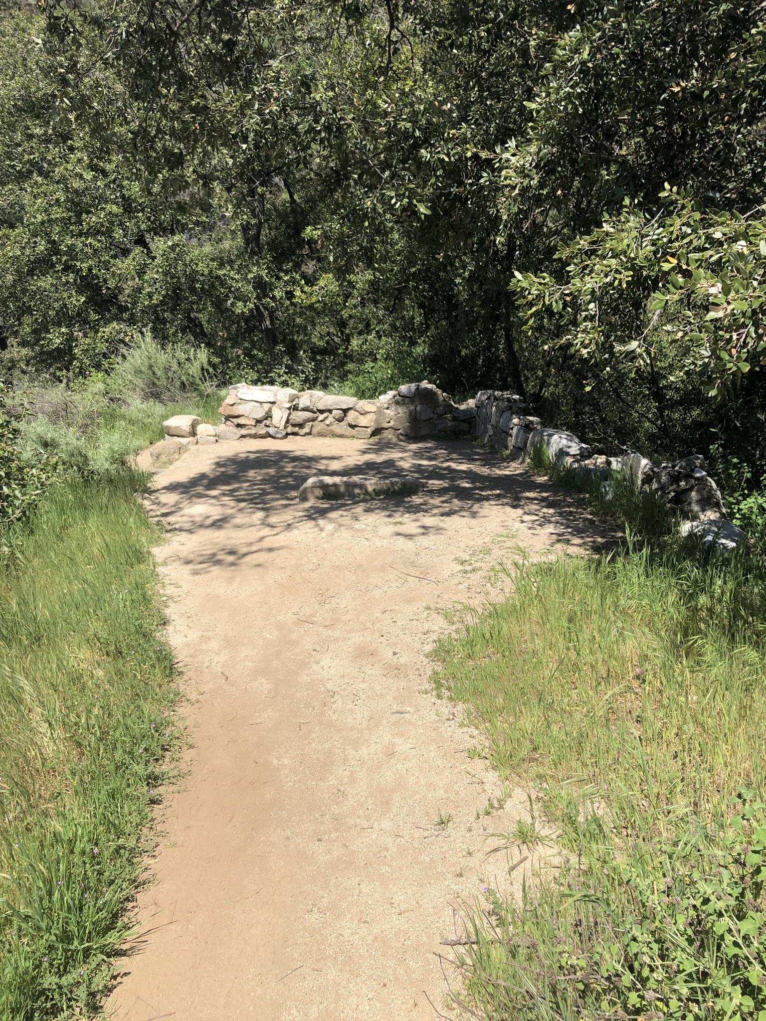 Sunny dirt path leads to a stone structure amidst green trees and grass.