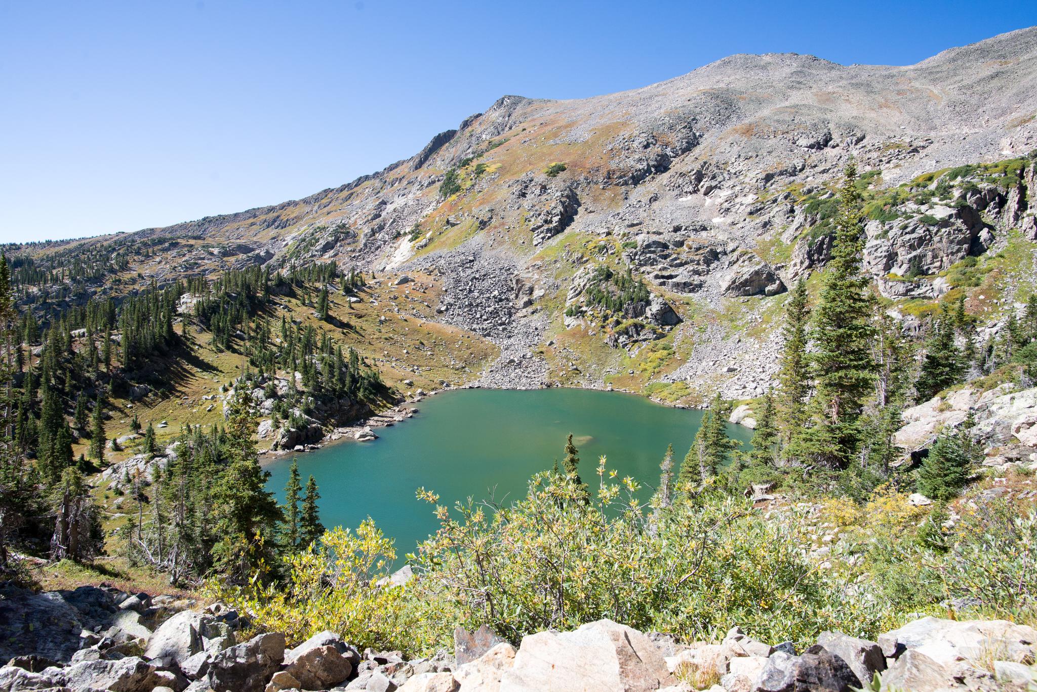 Bright turquoise lake in a rocky mountain basin with pine trees under clear sky.