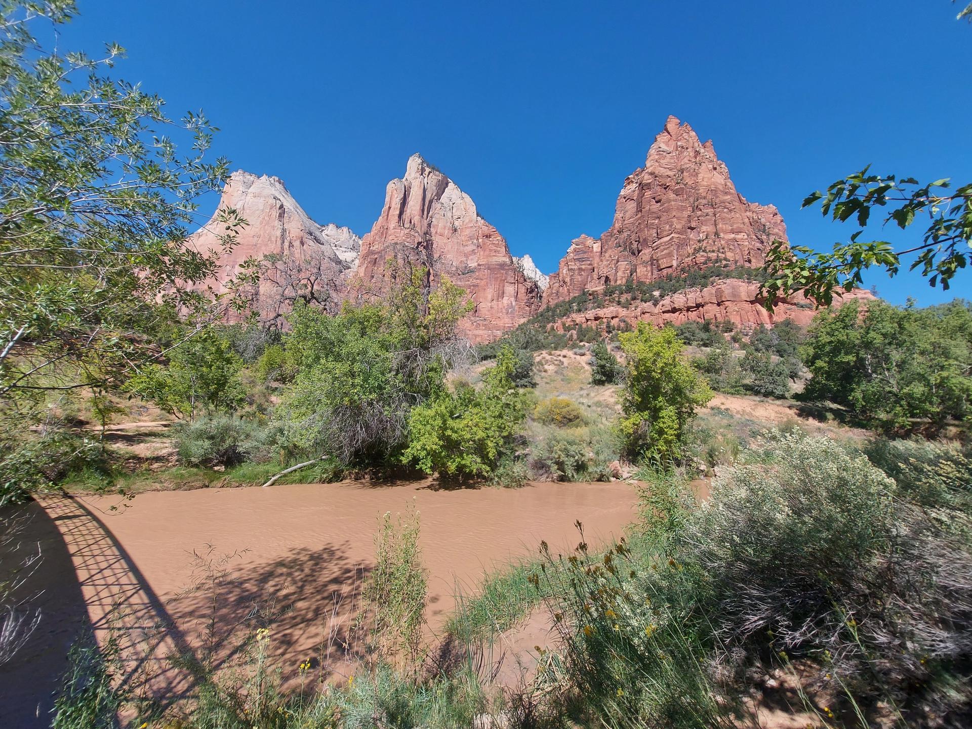 Towering red rock peaks, muddy river, and green trees under a clear blue sky.