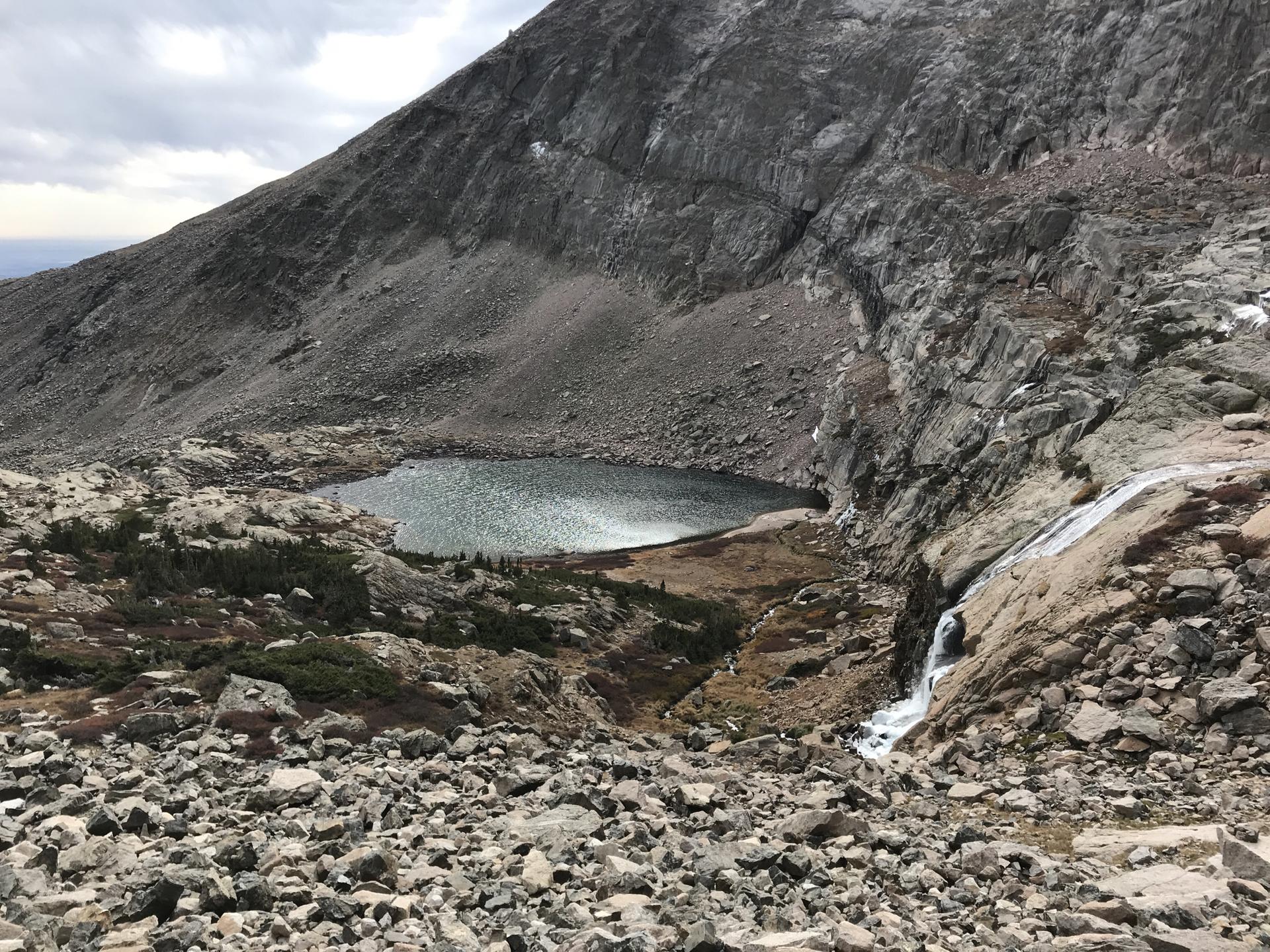 Overcast view of a small mountain lake fed by a waterfall amidst rocky terrain.