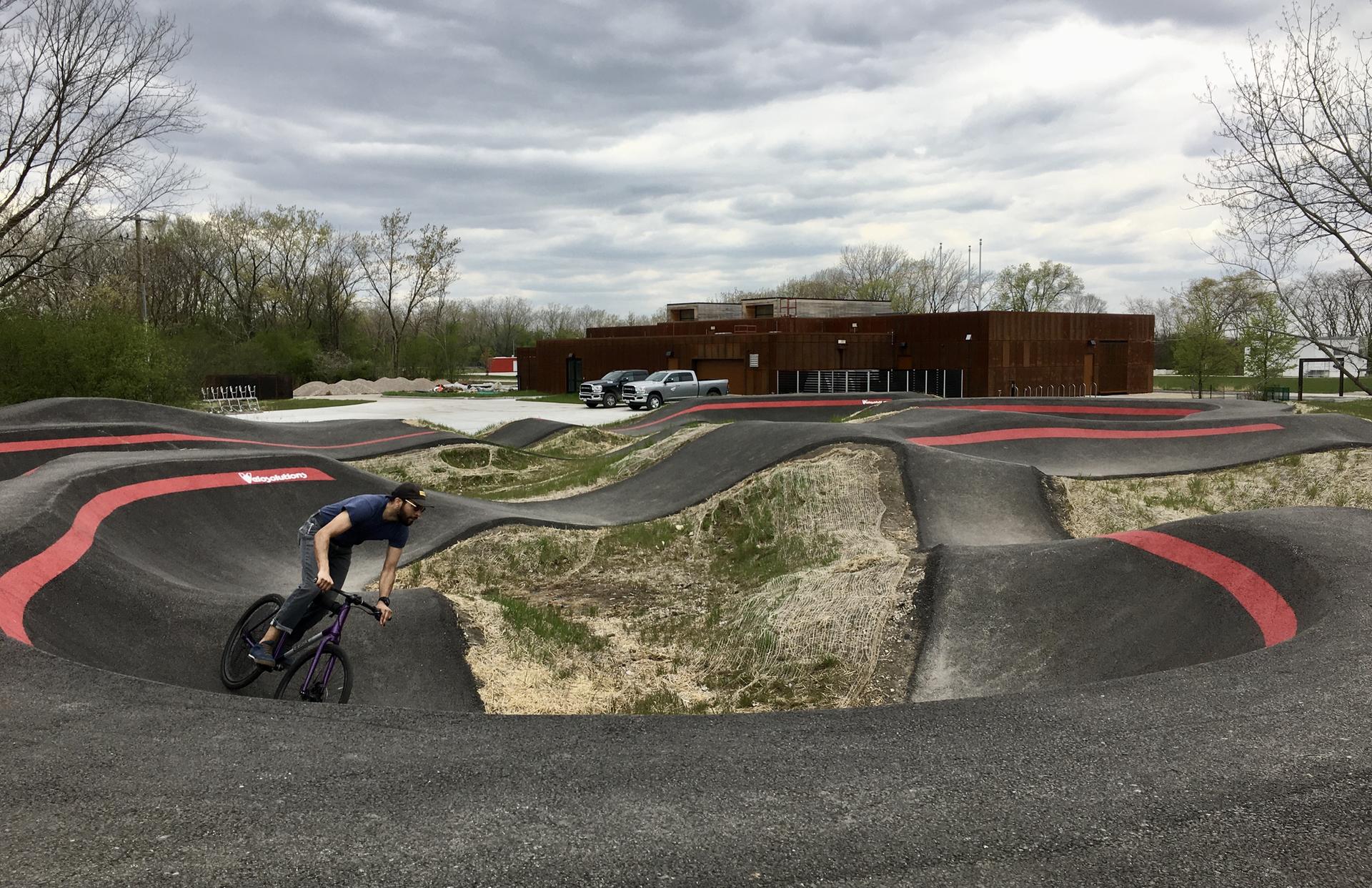 A person cycles on a dark pump track under an overcast sky.