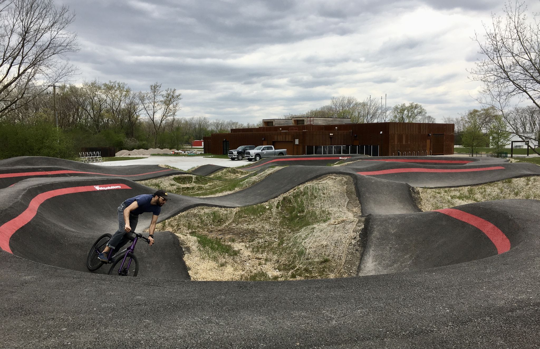 A person cycles on a dark pump track under an overcast sky. - Medium Terrain Trail