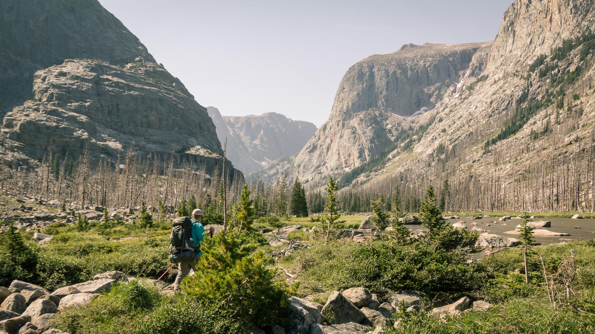 A hiker with a backpack and poles walks through a vast mountain valley.