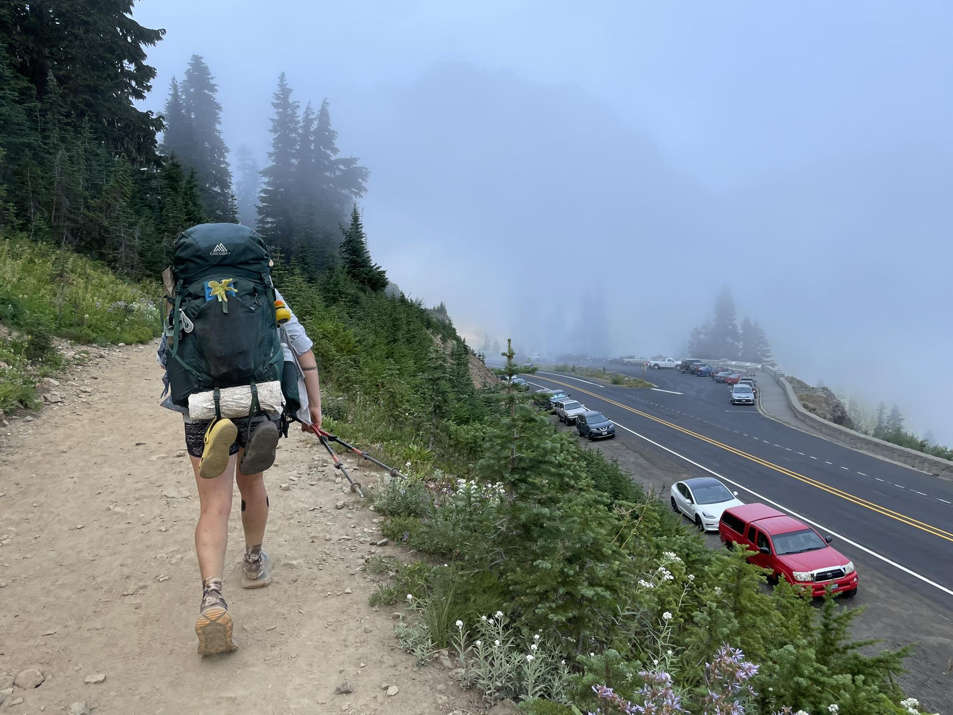 Hiker with large backpack walking on a foggy mountain path beside a road.