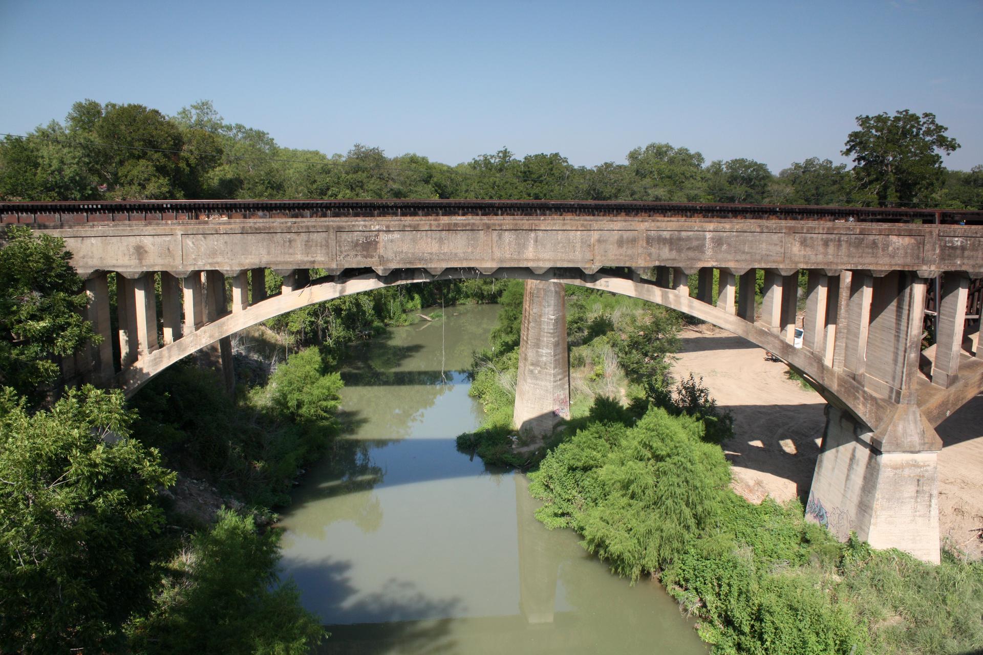 Large concrete arch bridge spans a green river under a clear blue sky.