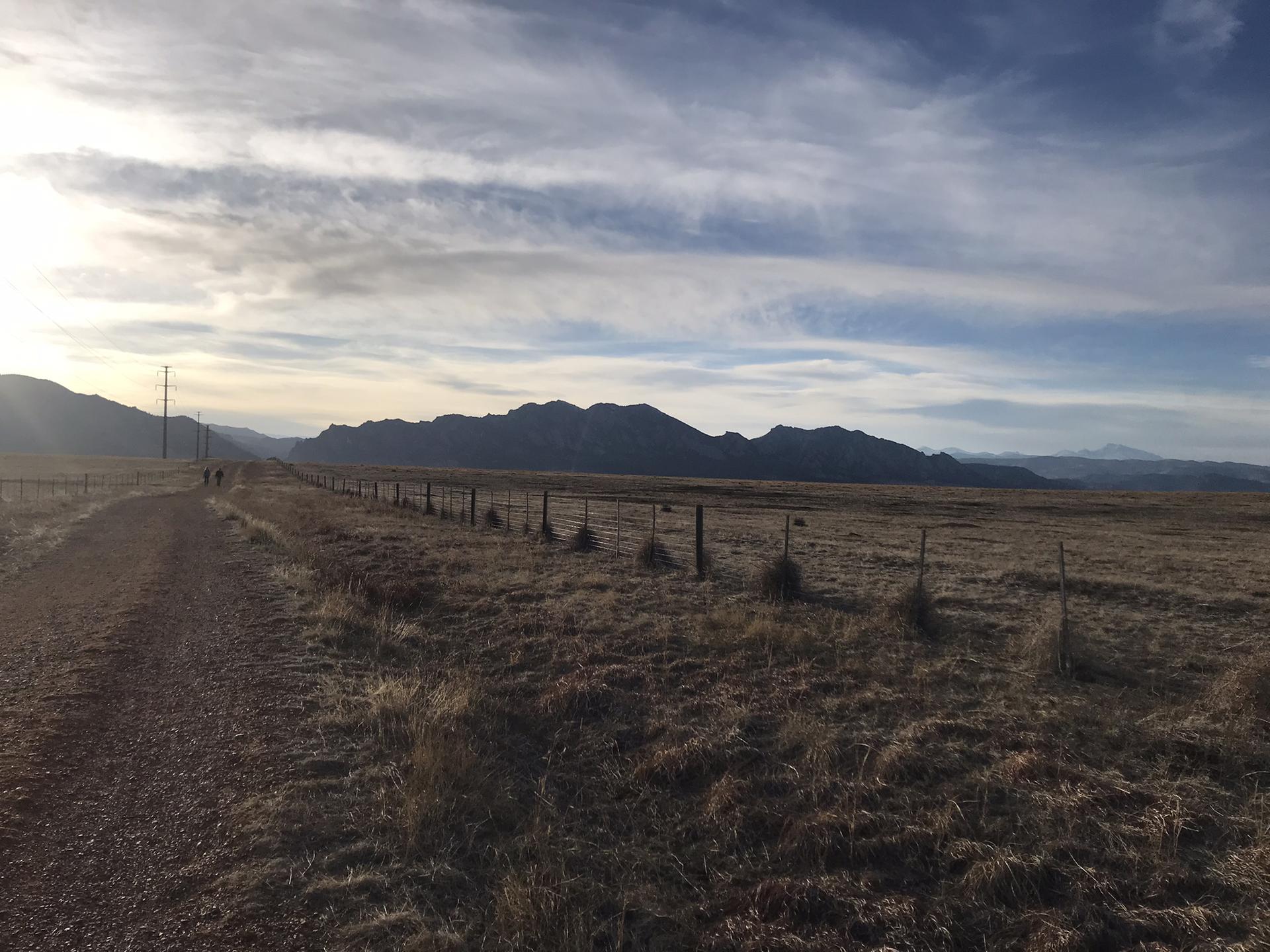 Dirt road beside a fence, leading towards distant mountains under a cloudy sky.