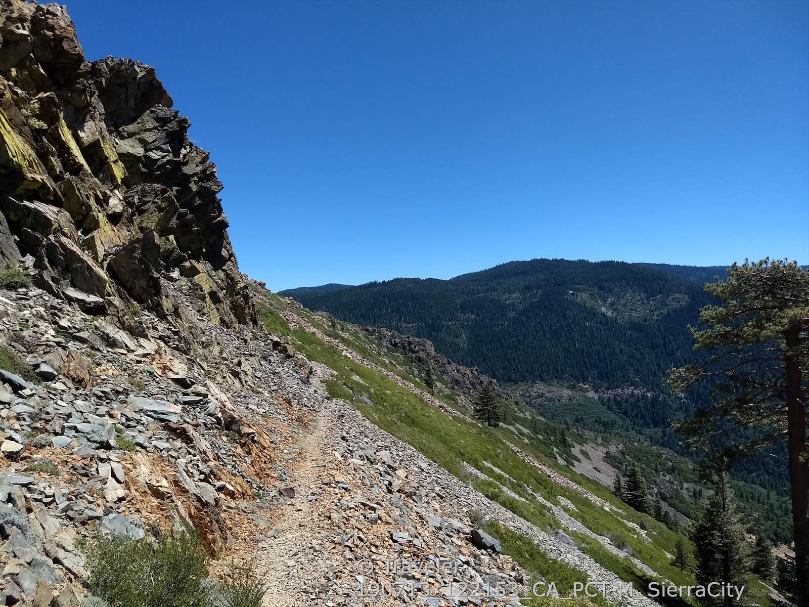 Narrow dirt trail on a steep, rocky mountainside under a clear blue sky.
