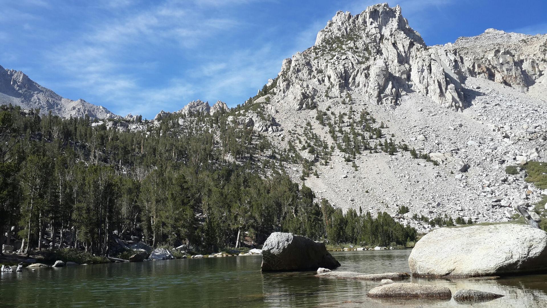 A clear lake with large rocks in the foreground, backed by a rugged mountain.