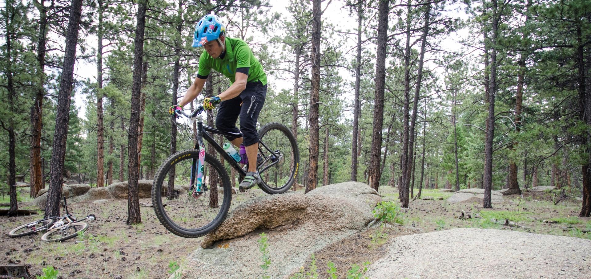 Mountain biker in green shirt rides over a large rock in a sunny forest.