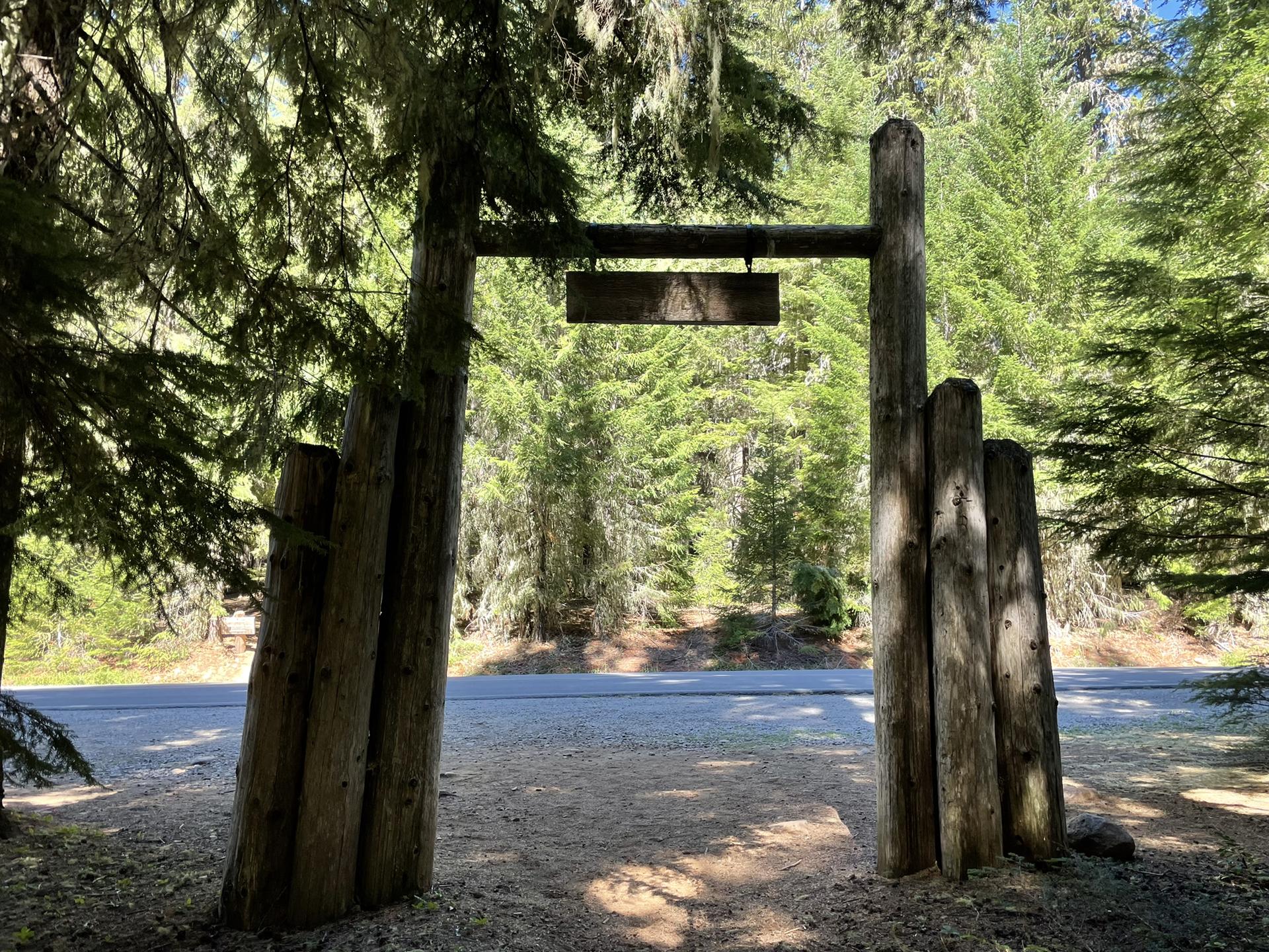Wooden archway entrance to a sunny forest path with a road beyond.