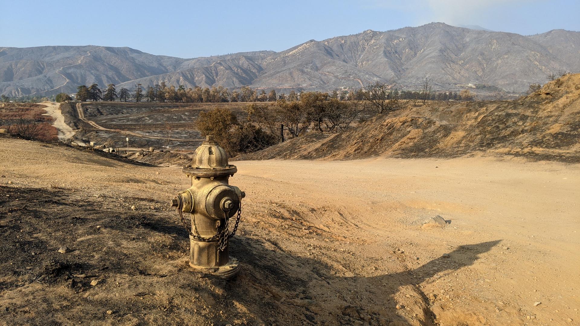 A fire hydrant stands in a dry, burnt landscape with mountains under bright daylight.