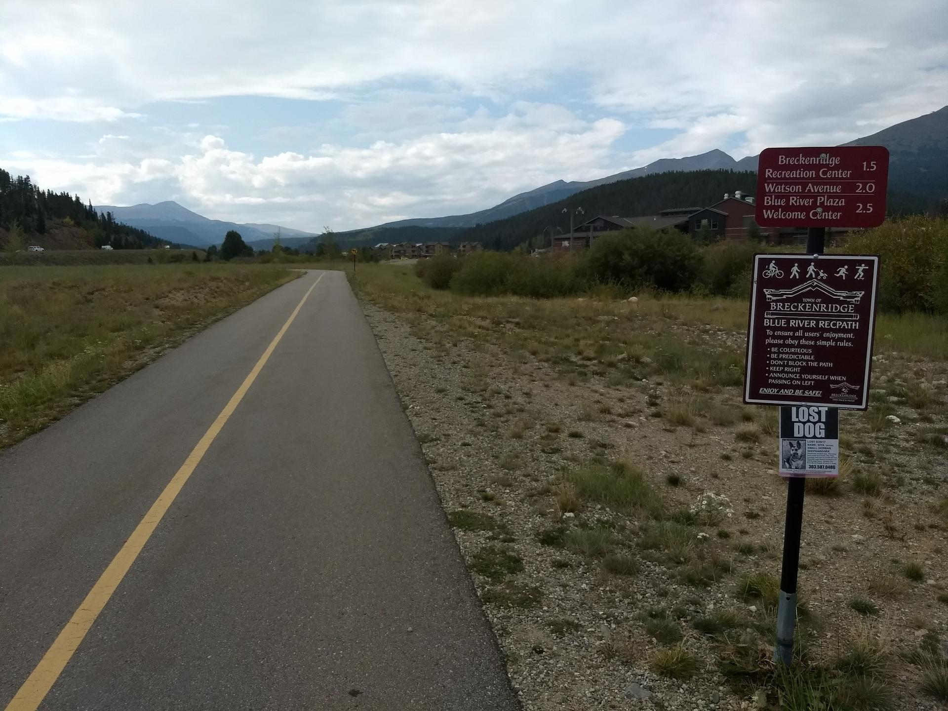 A paved path with signs, leading through grassy fields towards distant mountains.