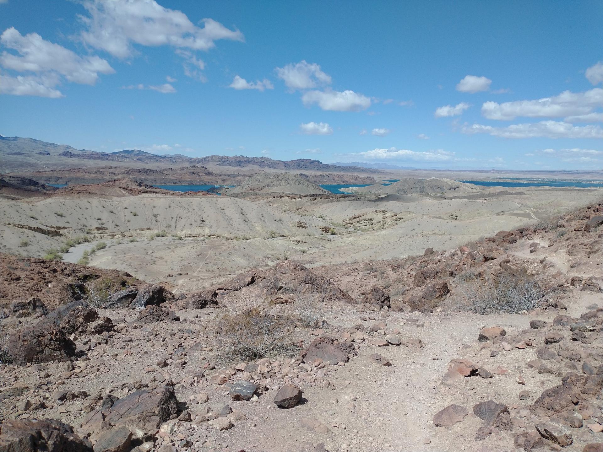 Vast desert landscape with rocky hills and distant blue water under a cloudy sky.