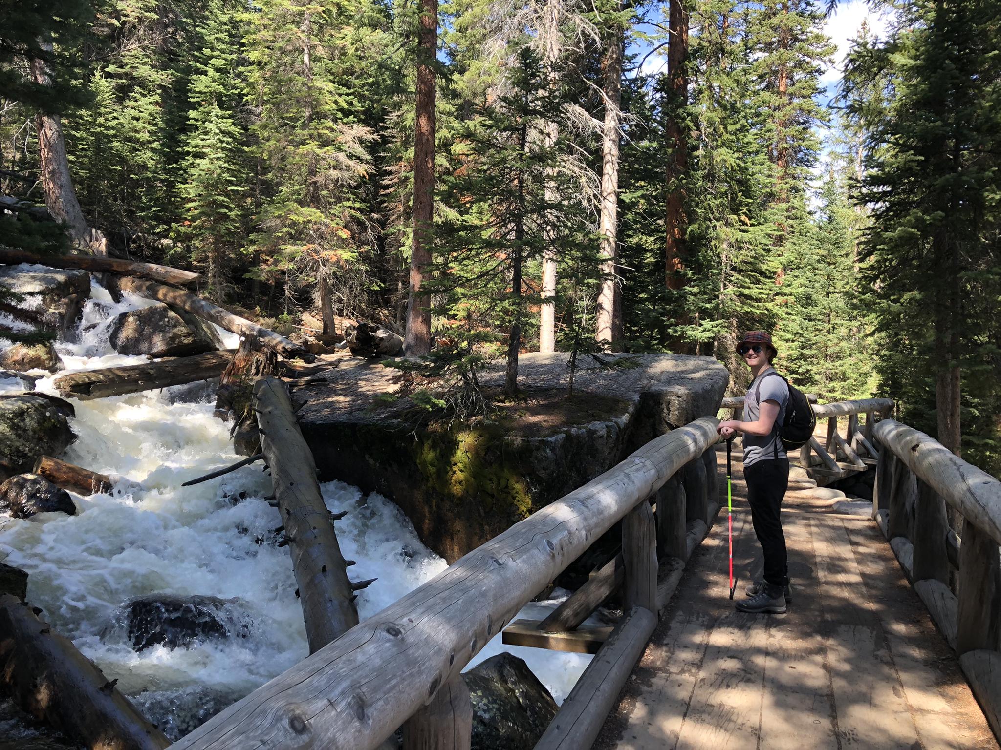 Hiker on a wooden bridge next to a rushing waterfall in a pine forest.