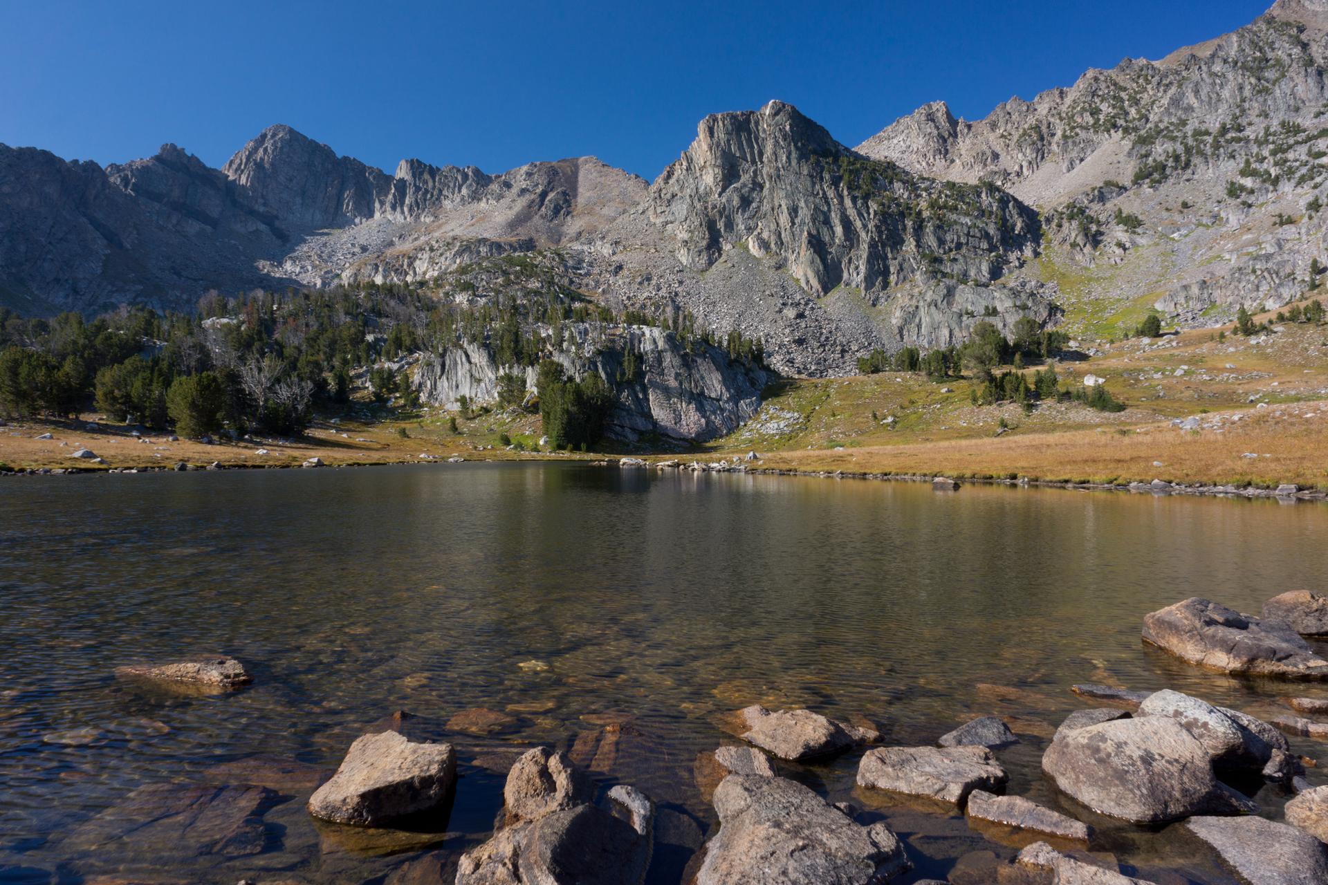 Clear mountain lake with rocky shore and towering peaks under blue sky.