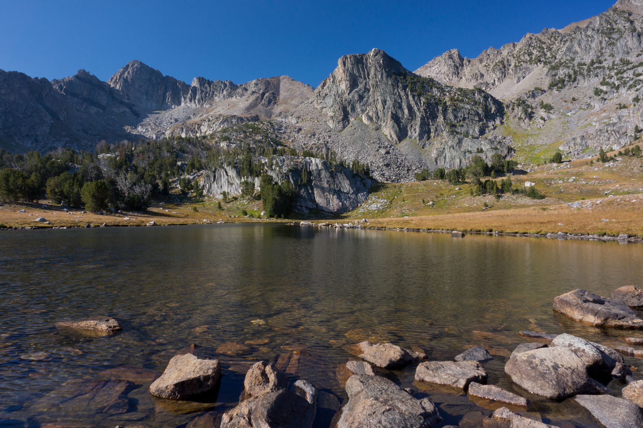 Clear mountain lake with rocky shore and towering peaks under blue sky. - Beehive Basin Trail