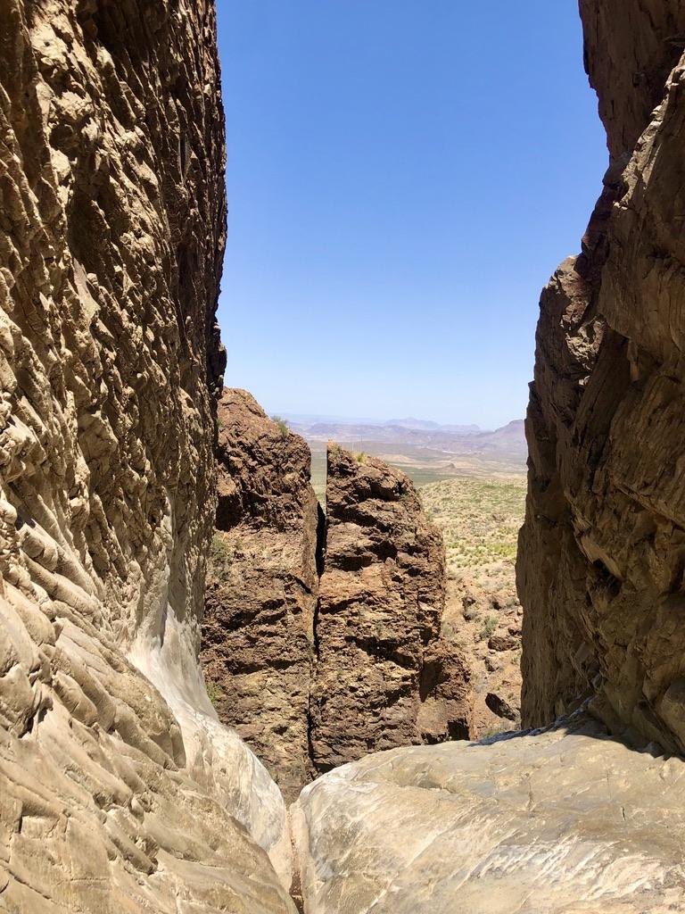 Narrow canyon opening reveals distant desert landscape under bright blue sky.