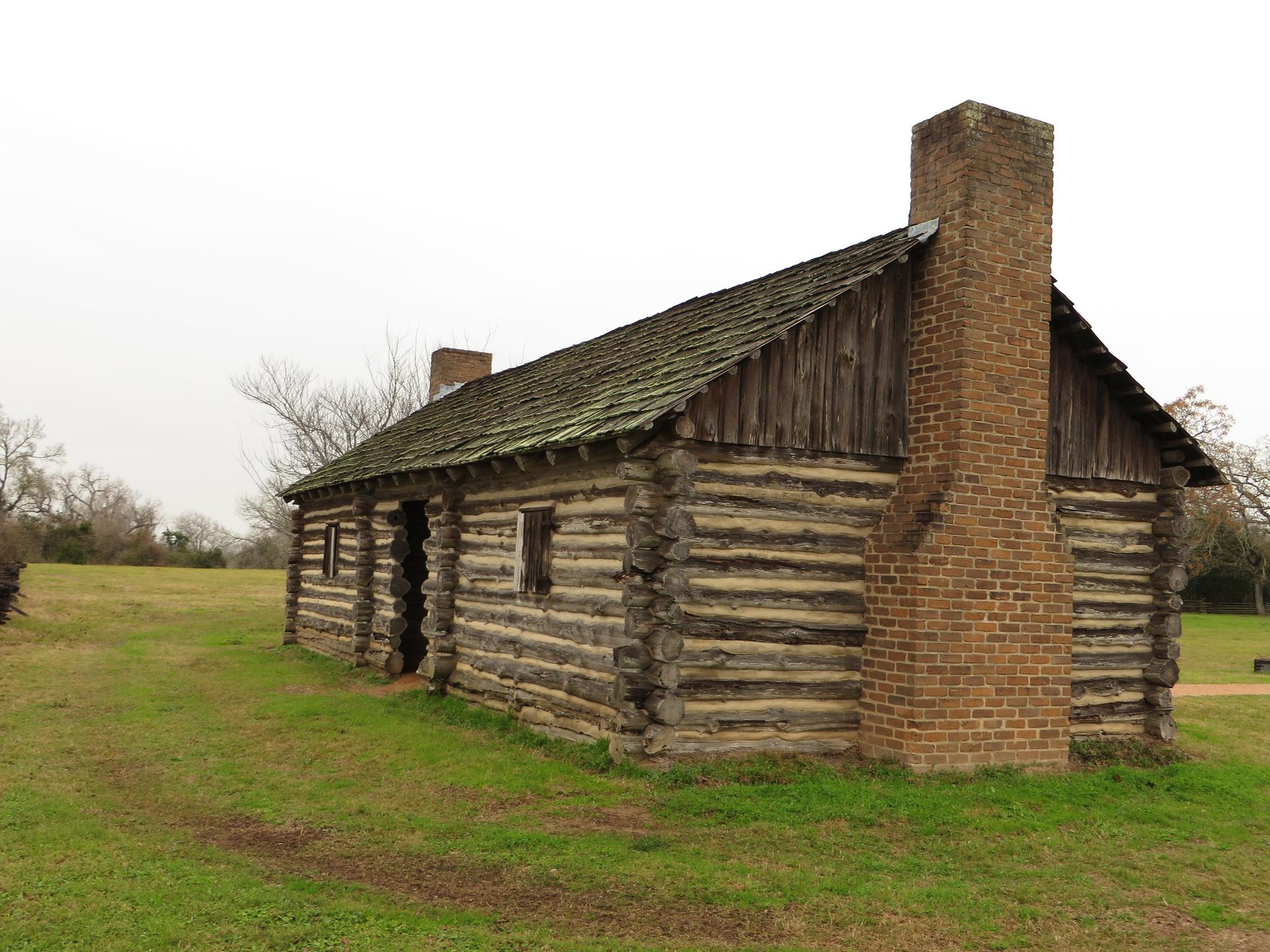 Old log cabin with a brick chimney in a grassy field under an overcast sky.