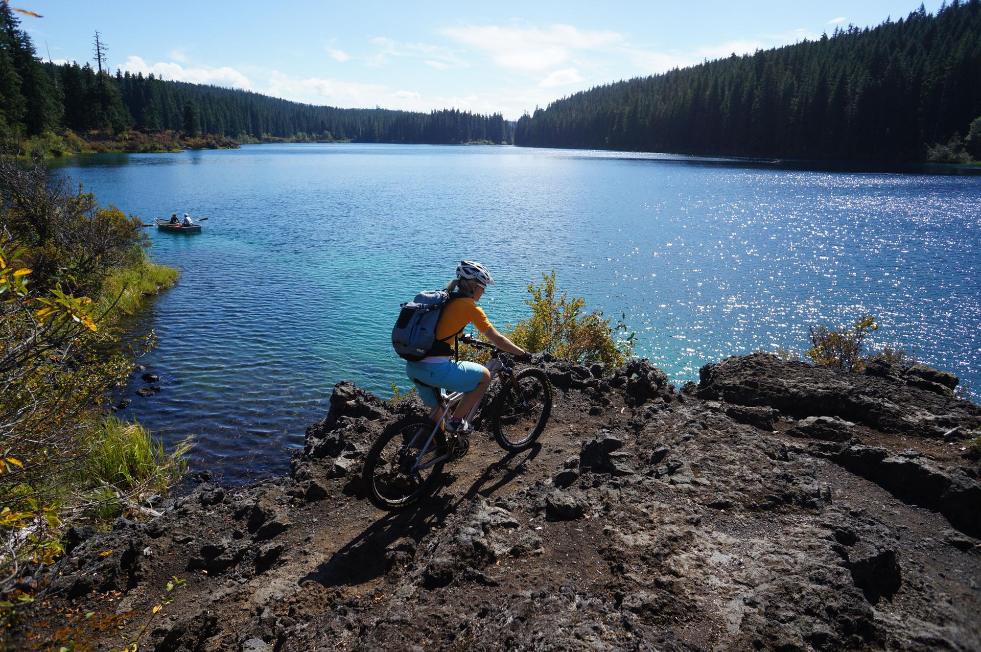 A person on a mountain bike overlooks a large, sparkling blue lake on a sunny day.