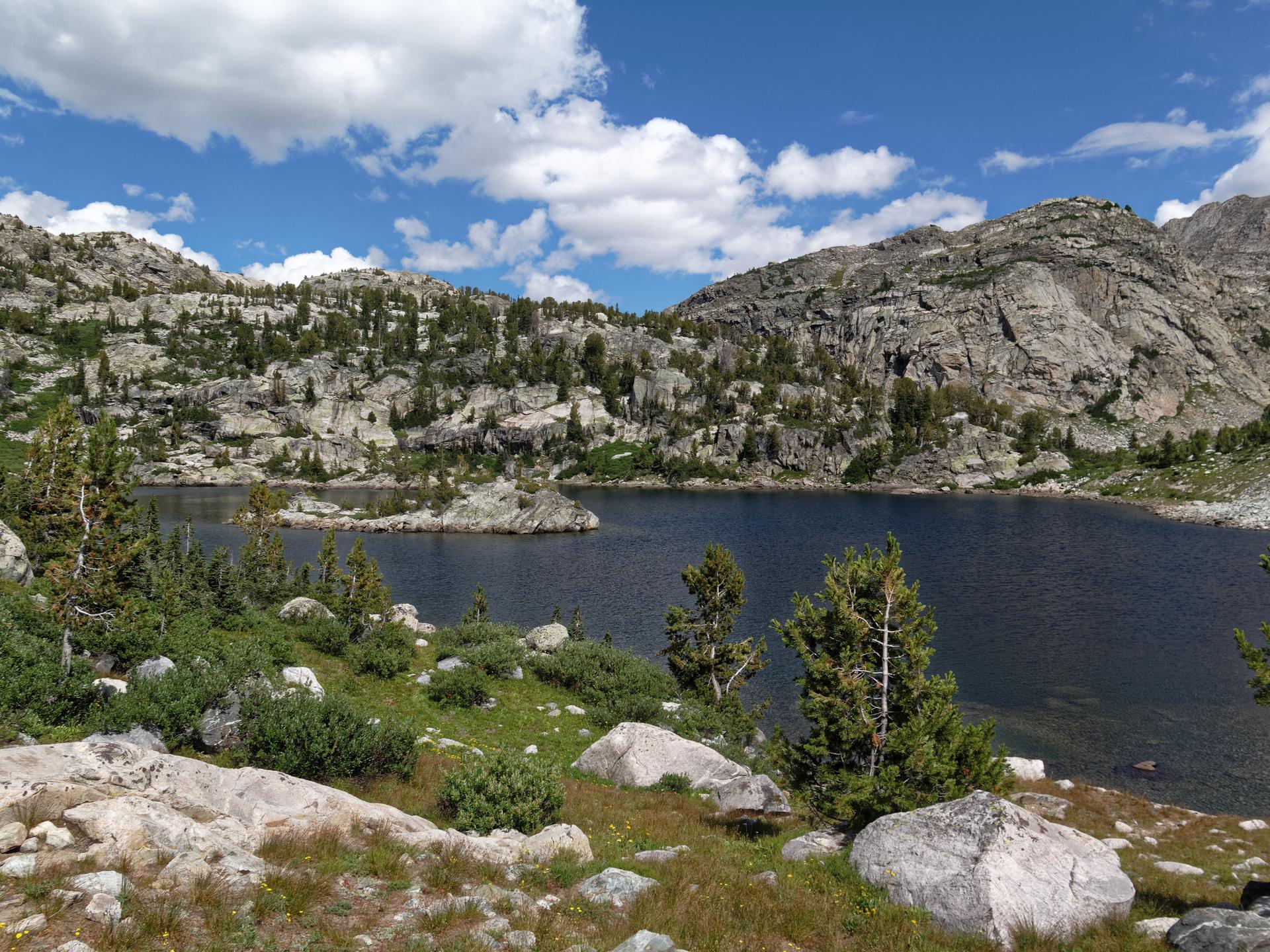 Lake with small rocky islands, surrounded by forested mountains, sunny.