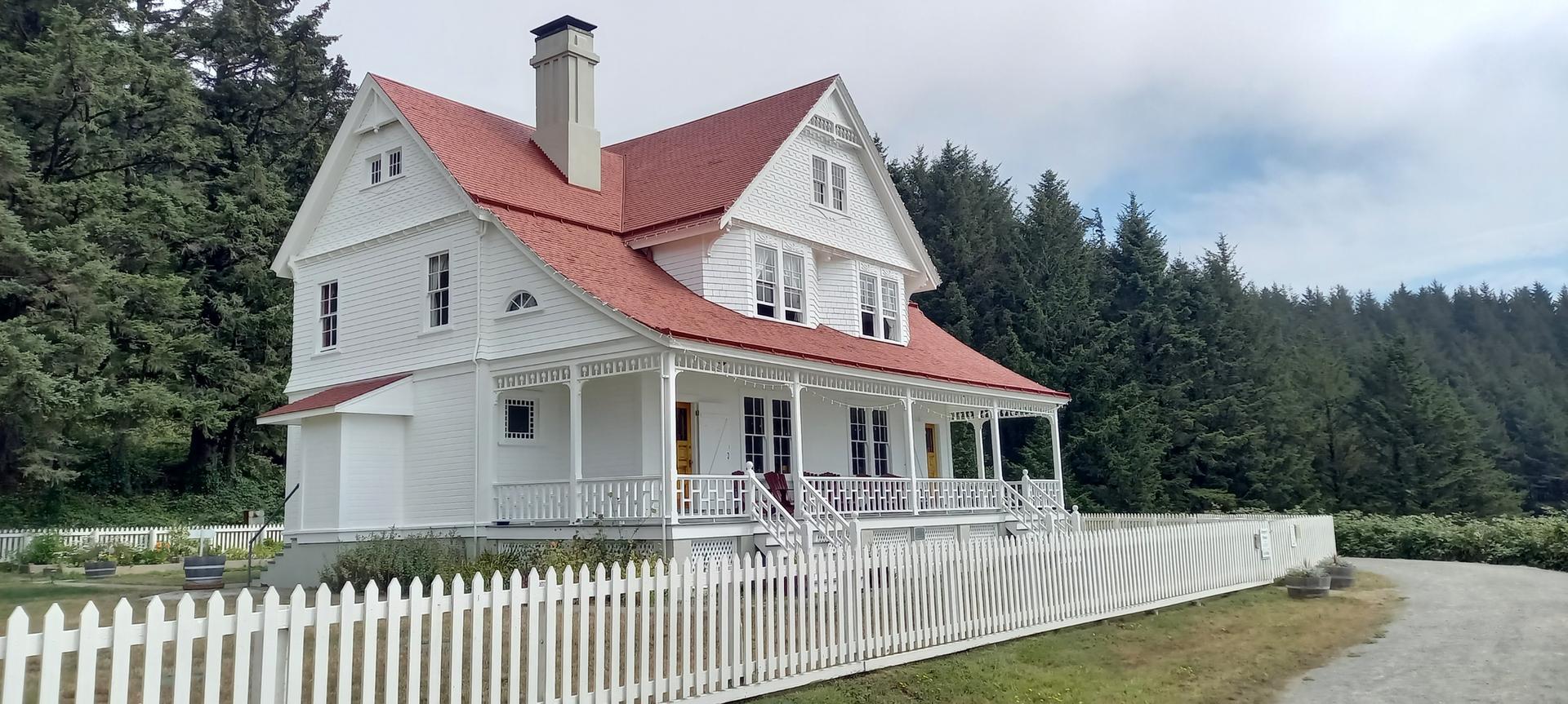 Large white house with a red roof and picket fence, surrounded by green forest.