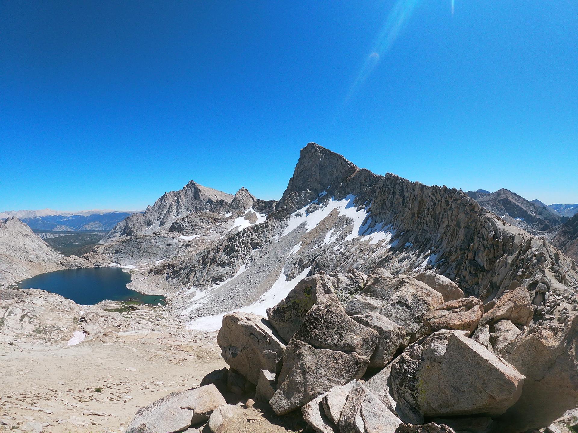 Rugged mountain peaks with snow patches overlook a distant lake under a bright sky.