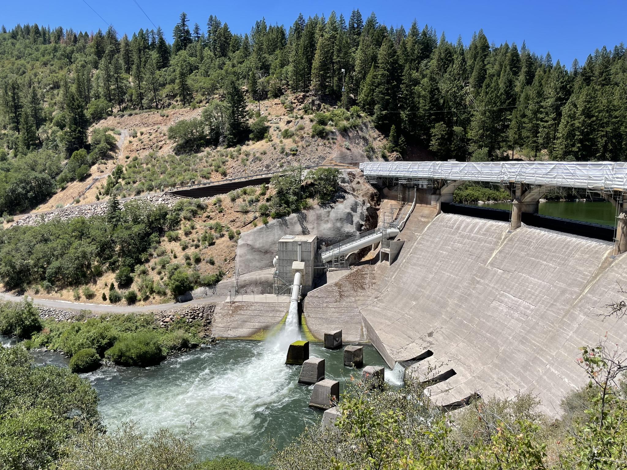 Powerful water discharge from a concrete dam, surrounded by forested hills.