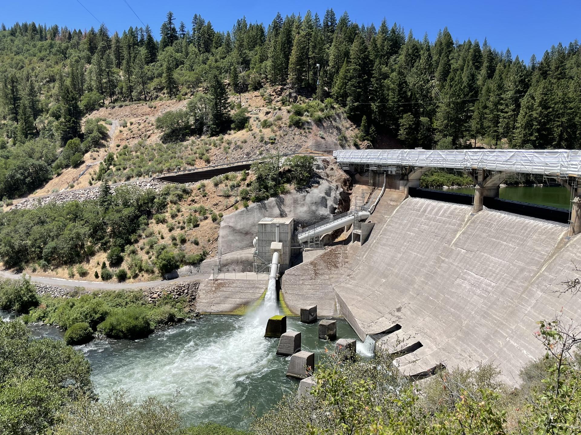 Powerful water discharge from a concrete dam, surrounded by forested hills.