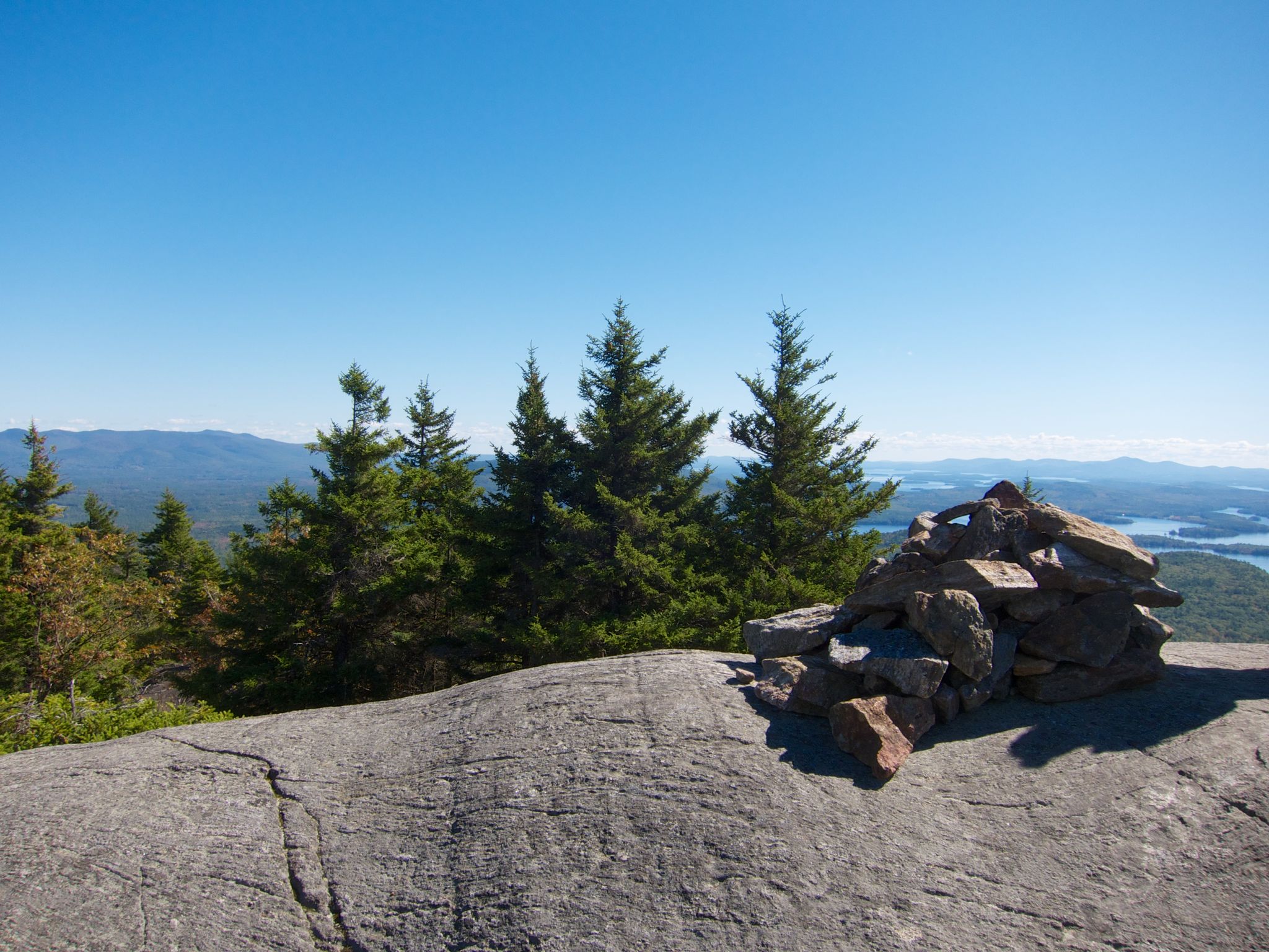 Rock cairn on a sunny, rocky summit with pine trees and distant mountains. - Mount Morgan and Mount Percival Loop