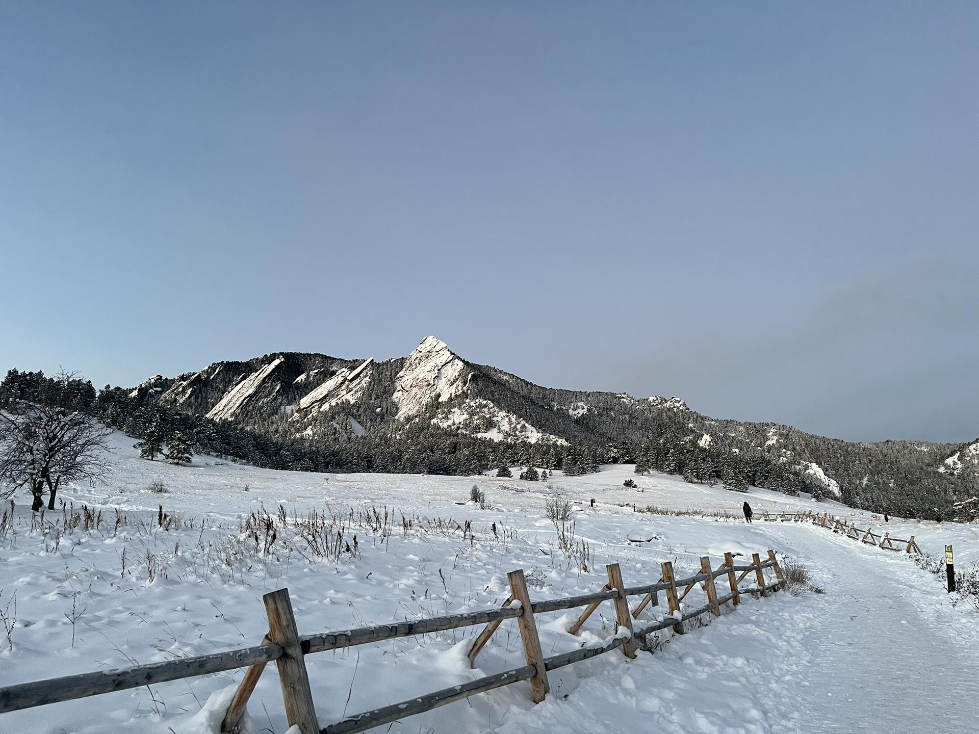 A snow-covered field with a wooden fence leads to snow-dusted mountains under a gray sky.