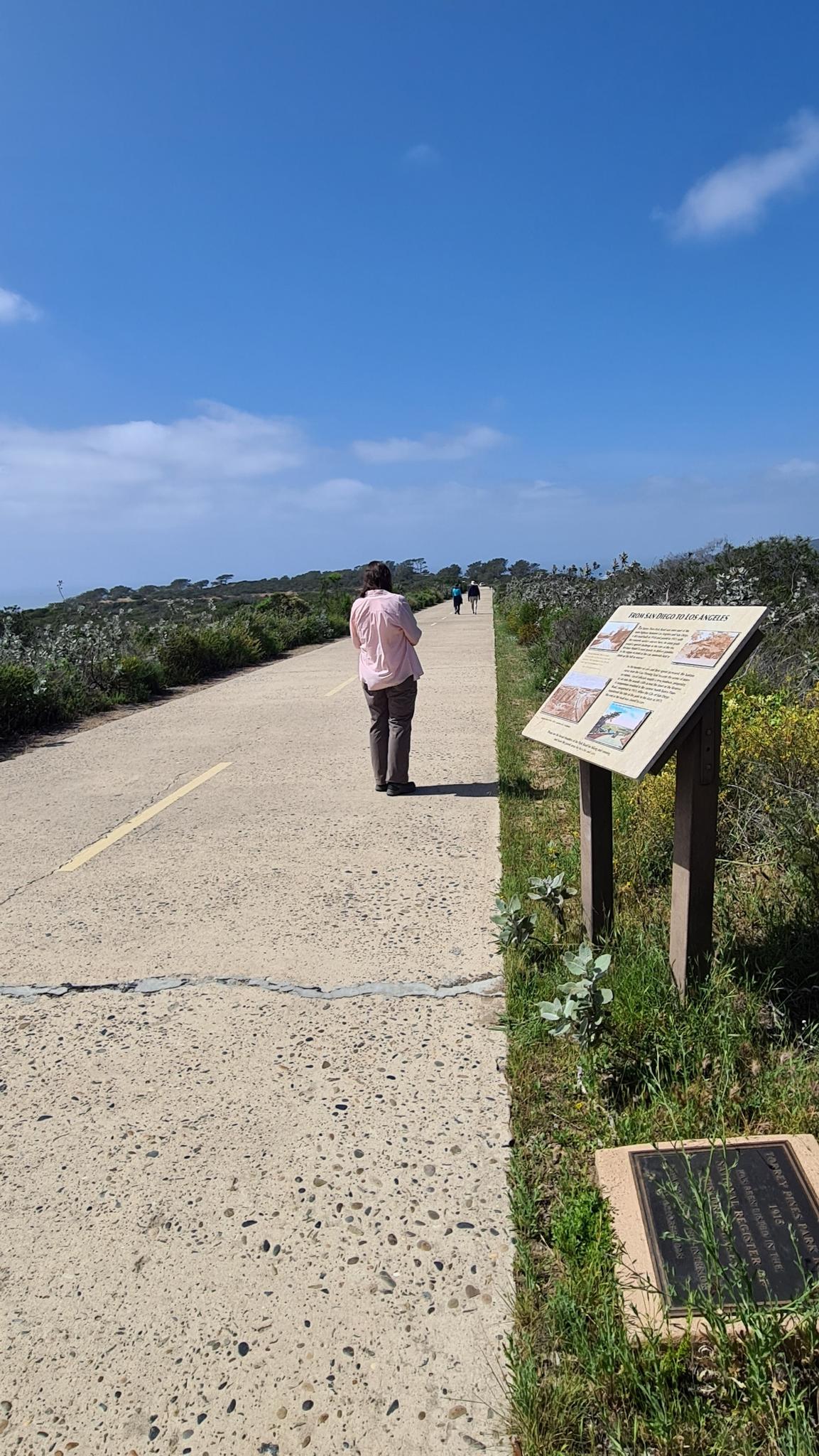 Person walking on a paved path next to an informational sign, with distant ocean.