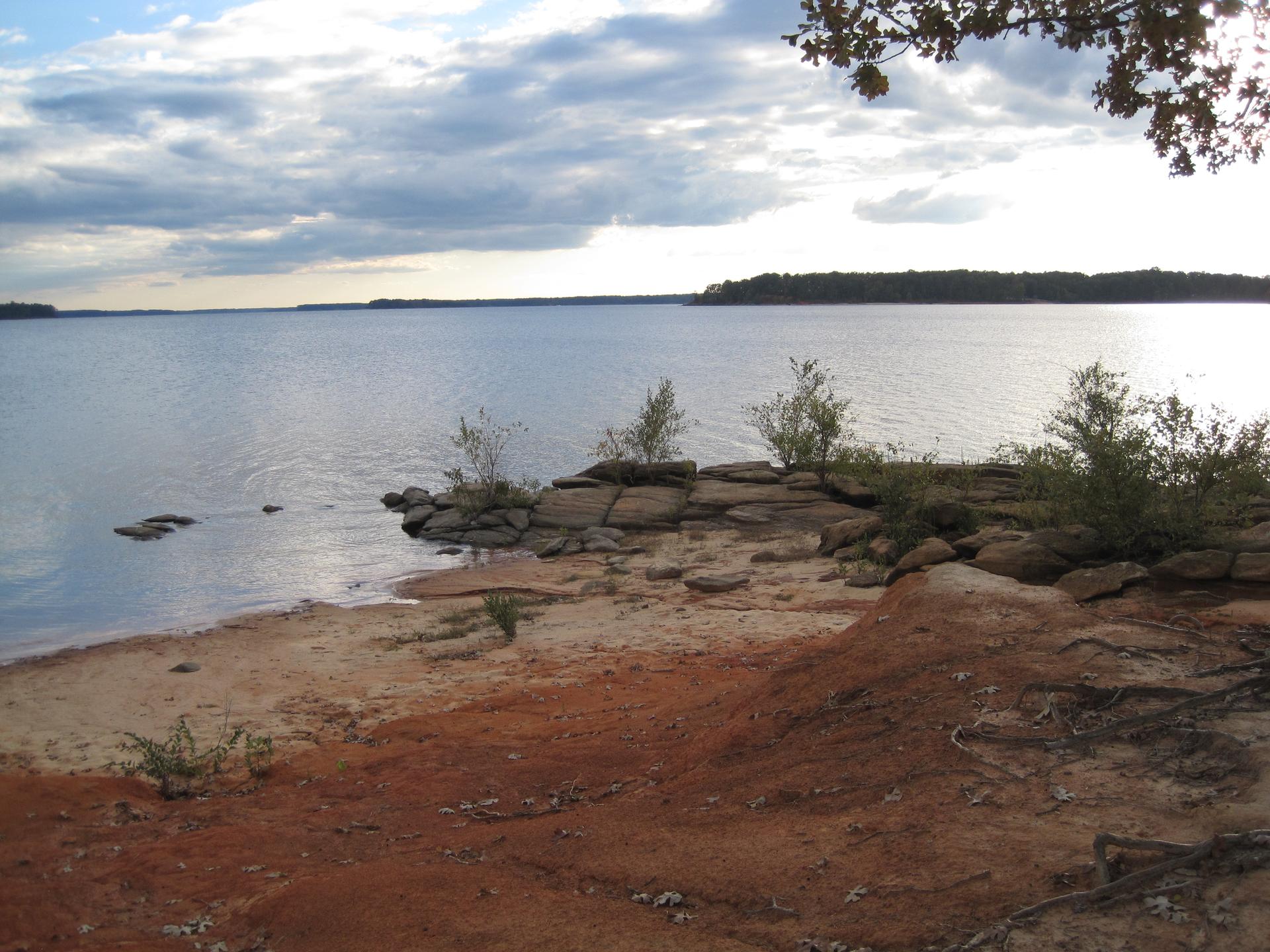 Wide lake view from a red sandy and rocky shore under cloudy skies.