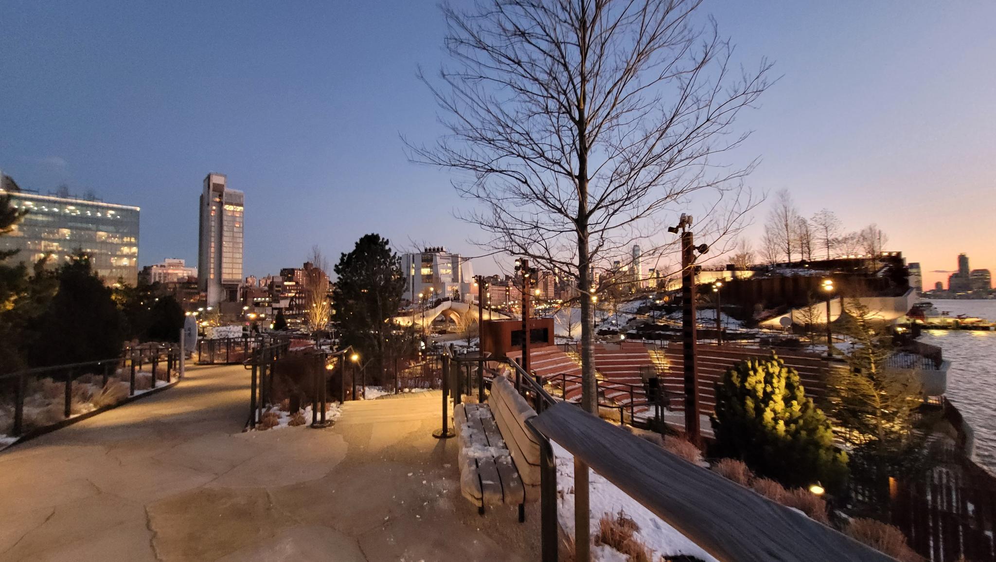 A park path with benches and bare trees, city skyline at dusk.