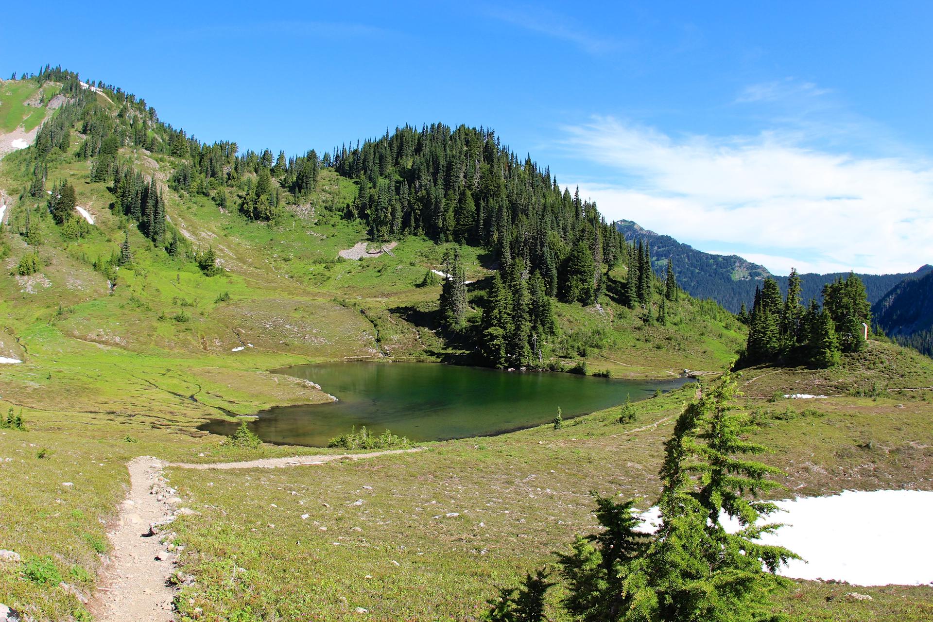 Green mountain landscape with a small lake, patches of snow, and blue sky.