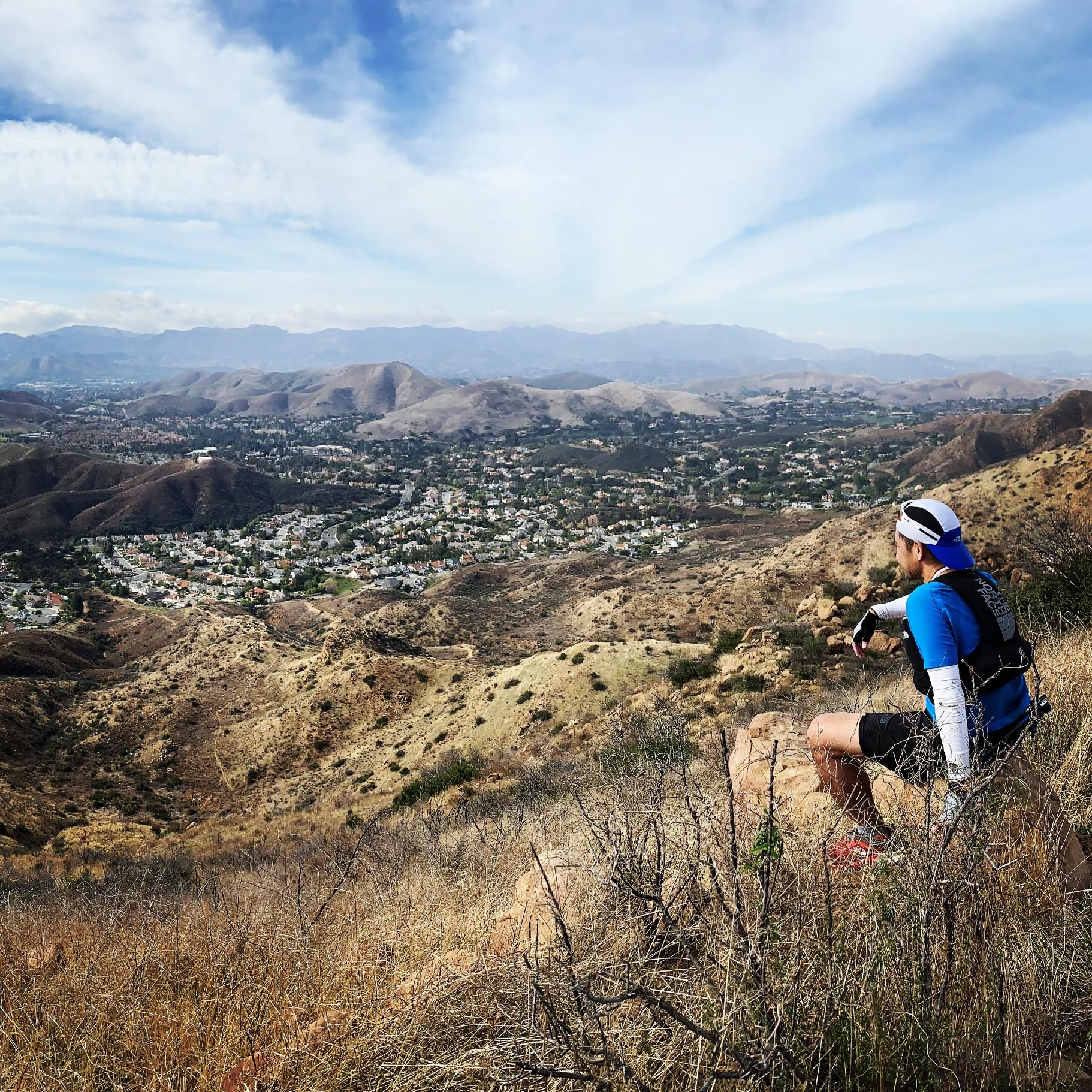 A person sits on a rock, overlooking a vast valley and distant town.