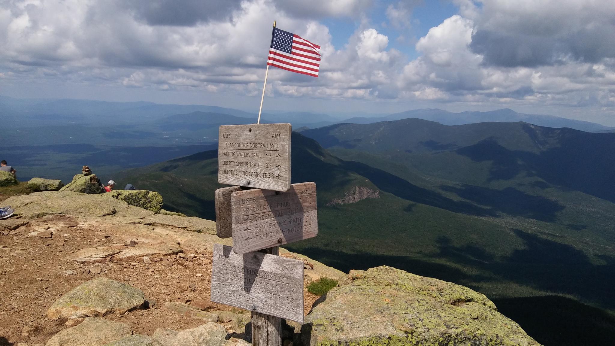 American flag on a wooden signpost on a rocky mountain peak, vast landscape.