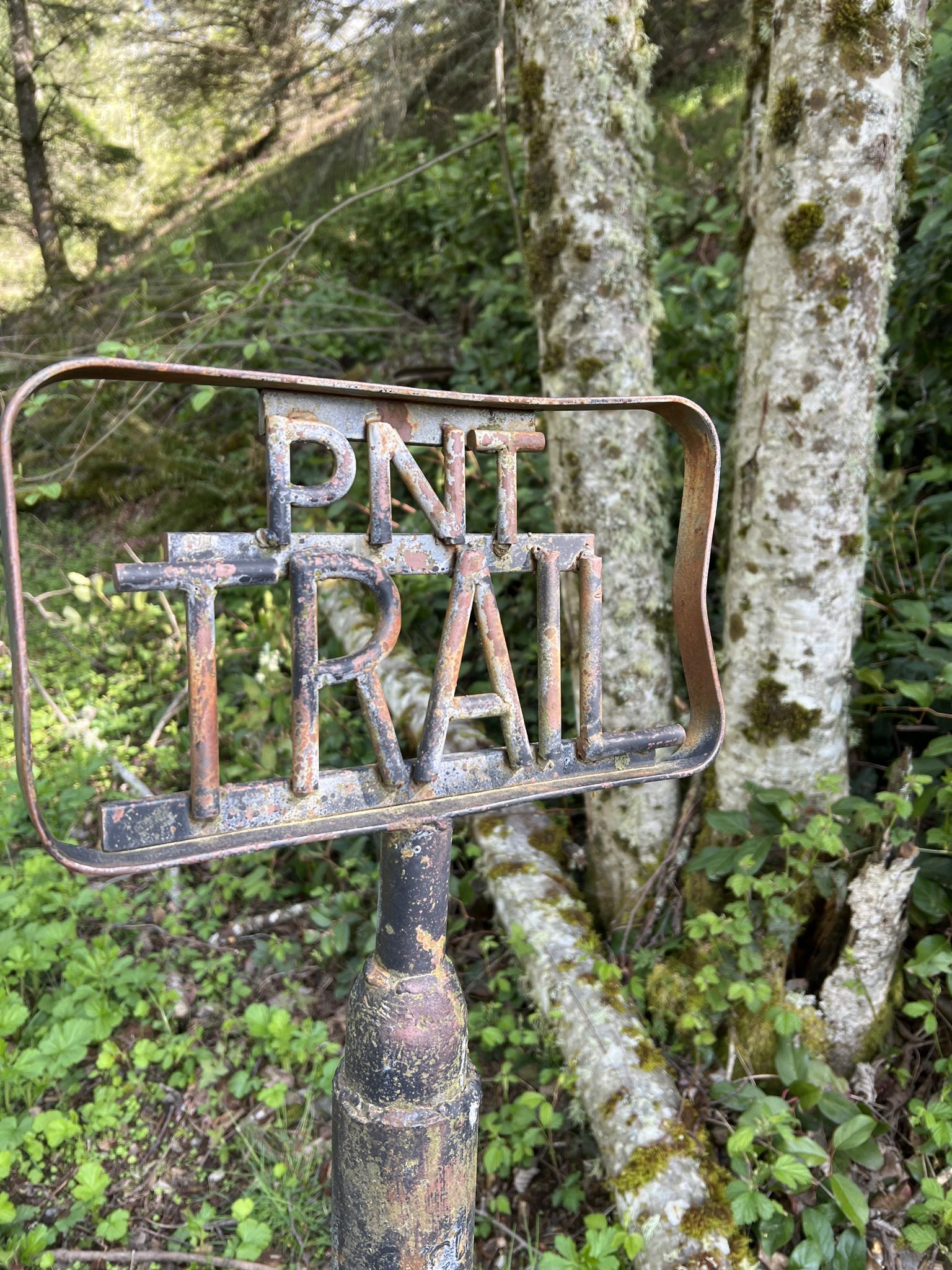 Close-up of a rustic metal trail sign in a green forest.
